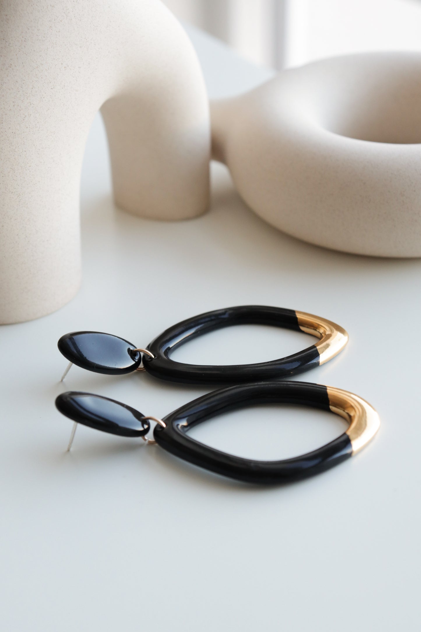 Rozenthal Jewelry’s Flow Earrings—a modern handmade black pair with gold tips—are displayed on a white surface with abstract beige ceramic sculptures in the background.