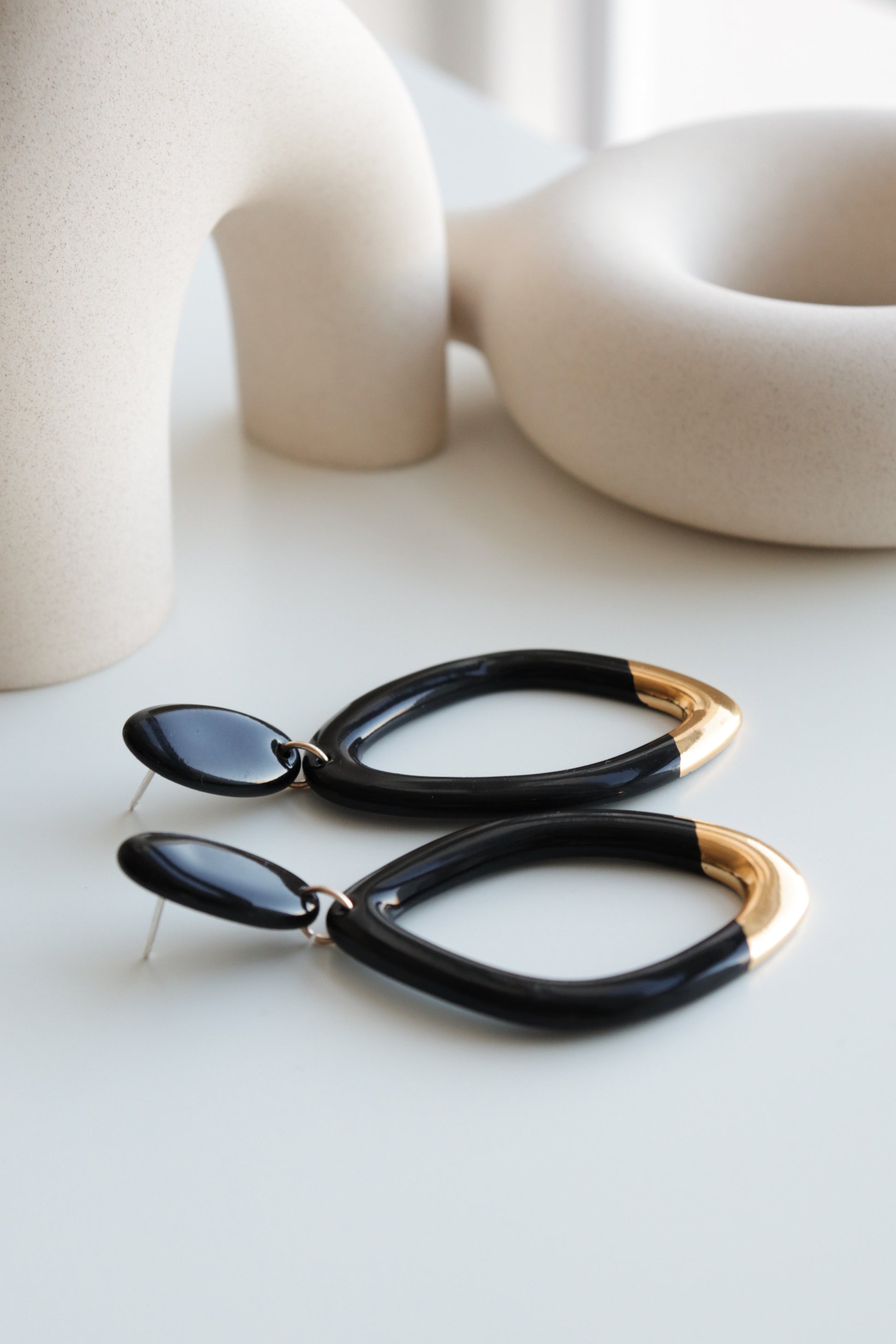 Rozenthal Jewelry’s Flow Earrings—a modern handmade black pair with gold tips—are displayed on a white surface with abstract beige ceramic sculptures in the background.
