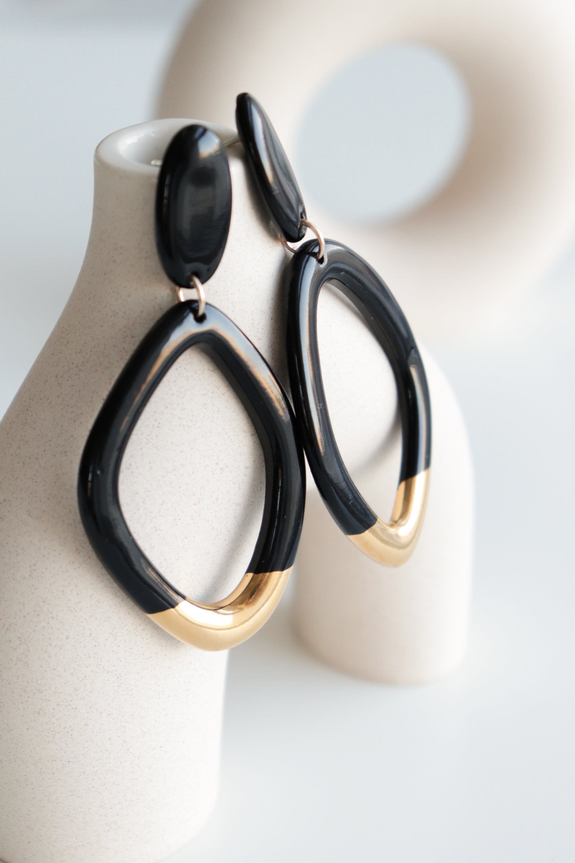 The Flow Earrings by Rozenthal Jewelry, modern black and gold geometric drops, hang on a white display stand. These standout designer earrings are hypoallergenic—perfect for sensitive ears—with a blurred circular decor piece behind.