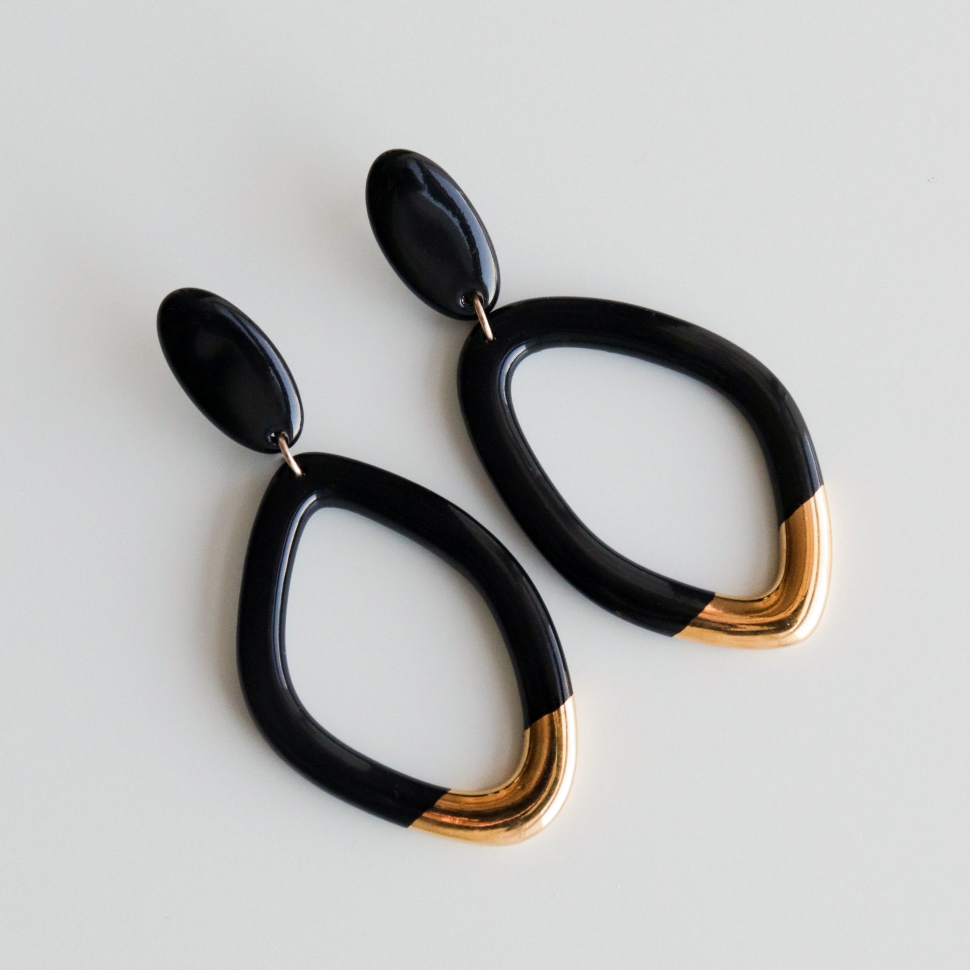 The Flow Earrings by Rozenthal Jewelry are handmade porcelain statement earrings from Latvia, featuring black teardrop loops with gold accents, shown on a plain white background.