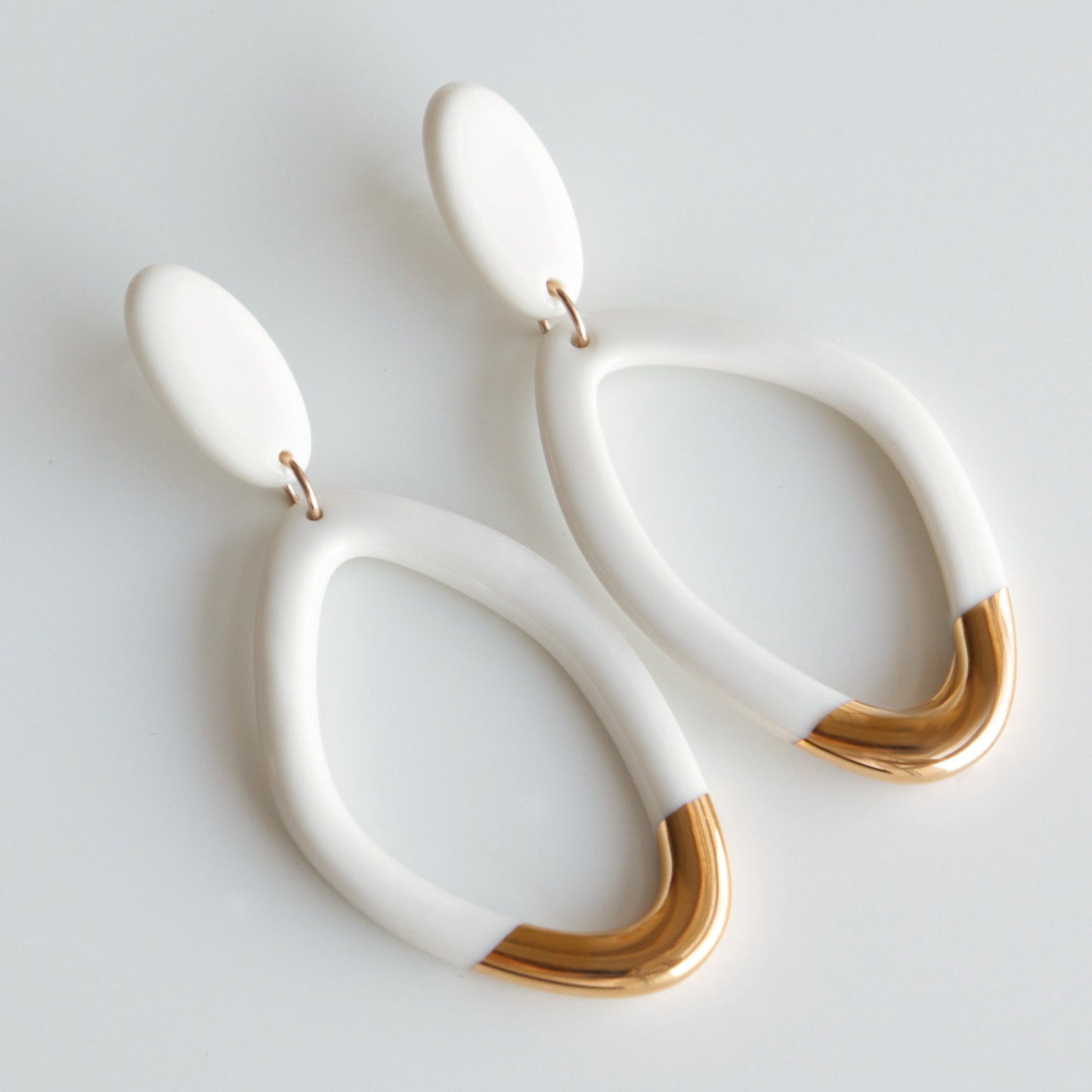 The Flow Earrings by Rozenthal Jewelry are modern porcelain statement earrings with white oval tops, large open loops, and gold metallic accents, handmade in Latvia and shown on a white background.