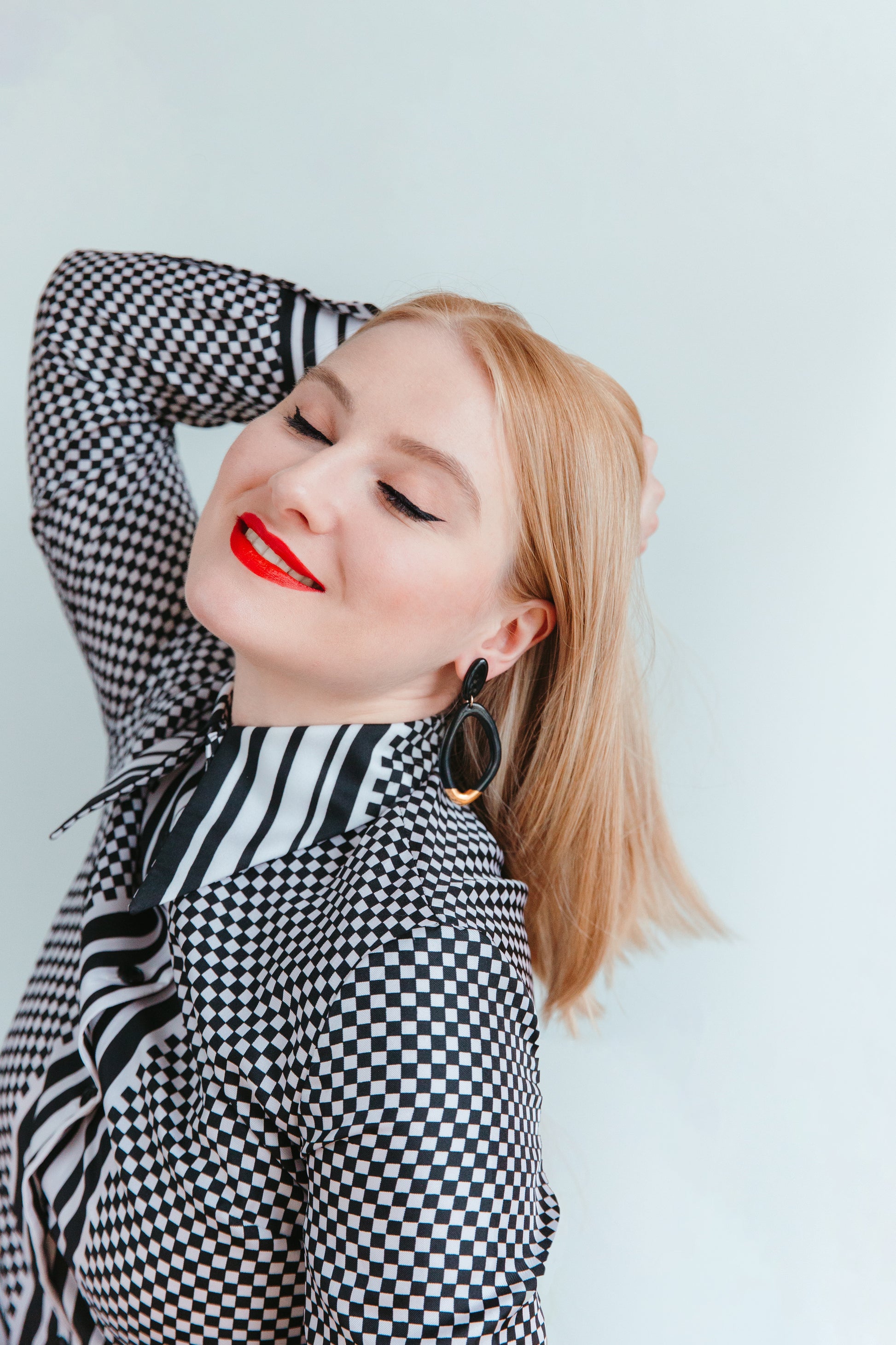 A smiling woman with red lipstick poses with one hand in her blonde hair, wearing Rozenthal Jewelrys lightweight, waterproof Flow Earrings and a black-and-white checkered blouse against a light background.