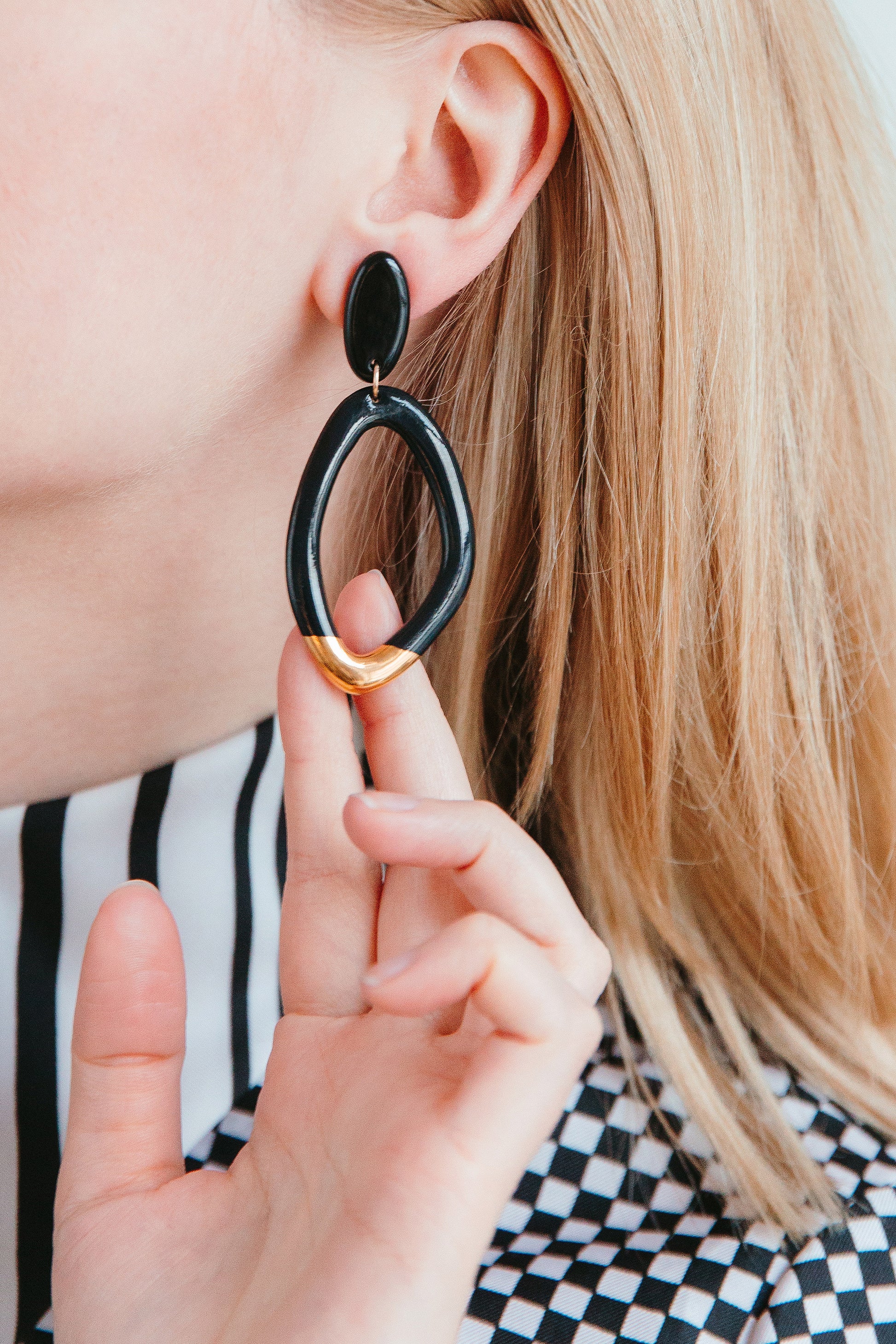 A person with blonde hair wears a black and white patterned top while holding a large, oval-shaped Flow Earring by Rozenthal Jewelry—handmade porcelain with gold accents from Latvia—near their ear against a blurred background.
