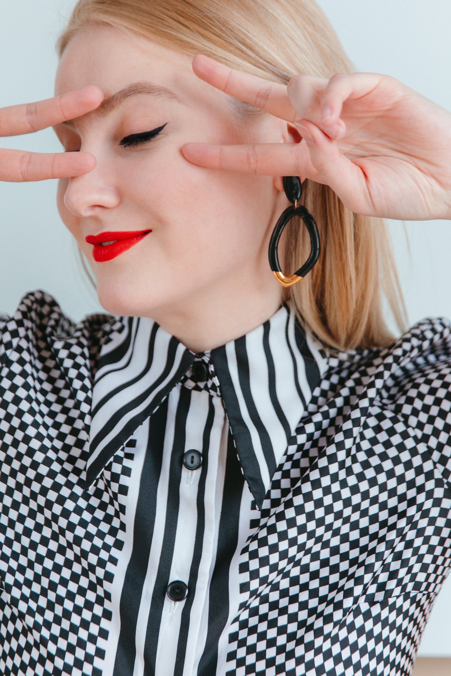A woman with long blonde hair, red lipstick, and winged eyeliner flashes peace signs by her eyes. She wears a black and white patterned shirt and showcases Rozenthal Jewelry’s handmade Flow Earrings, highlighting her love for sustainable accessories.