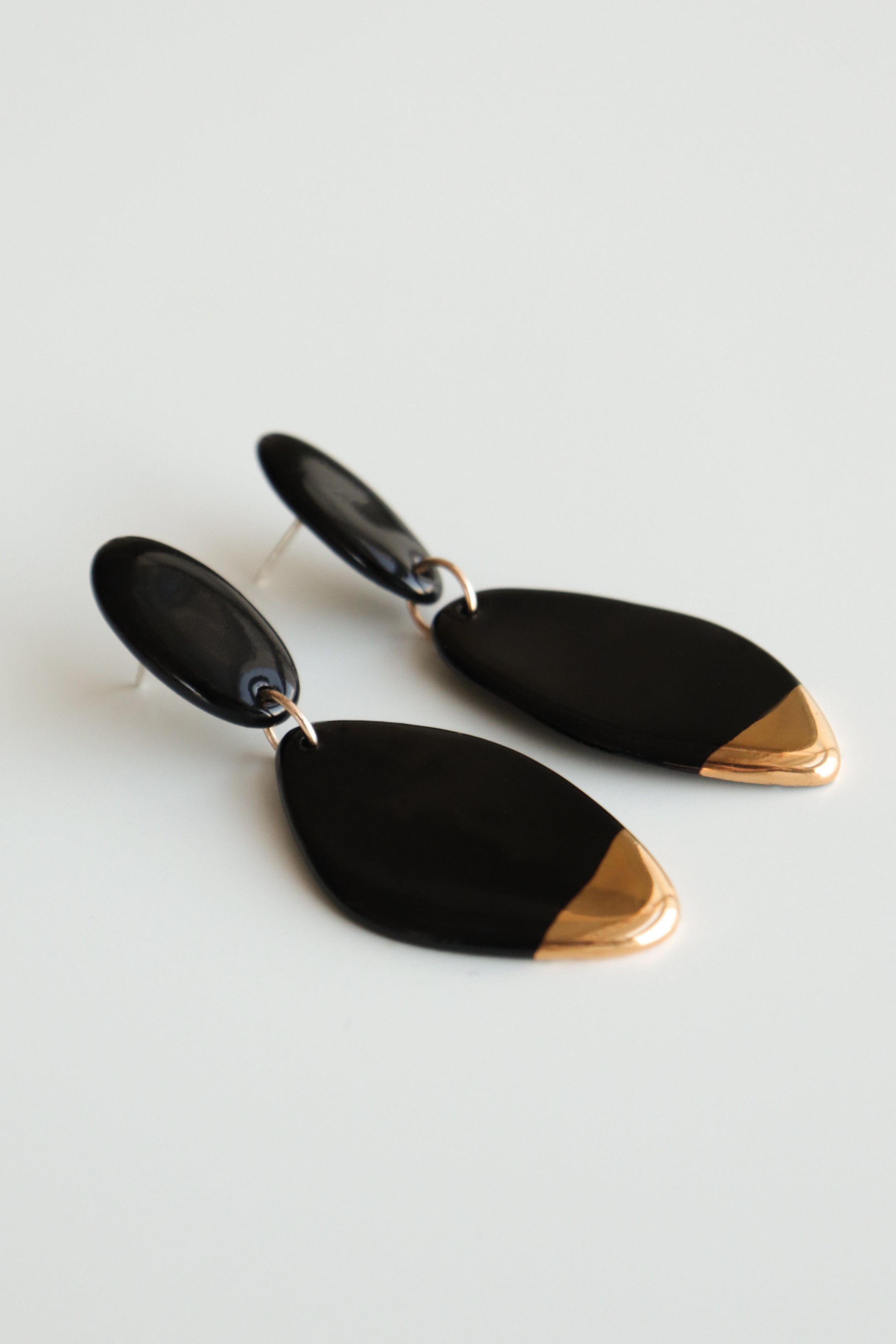 The Flow Earrings by Rozenthal Jewelry are handmade black porcelain statement pieces with gold tips, featuring a shiny finish and elongated oval shape. Made in Latvia, they are showcased on a white background.