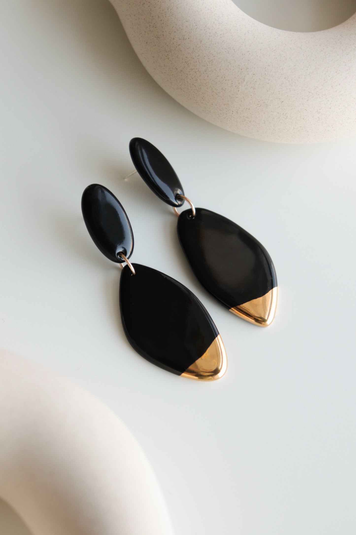 Flow Earrings by Rozenthal Jewelry—modern black drop earrings with gold tips, handmade in Latvia. Shown on a soft white surface with curved beige decor accents in the background.