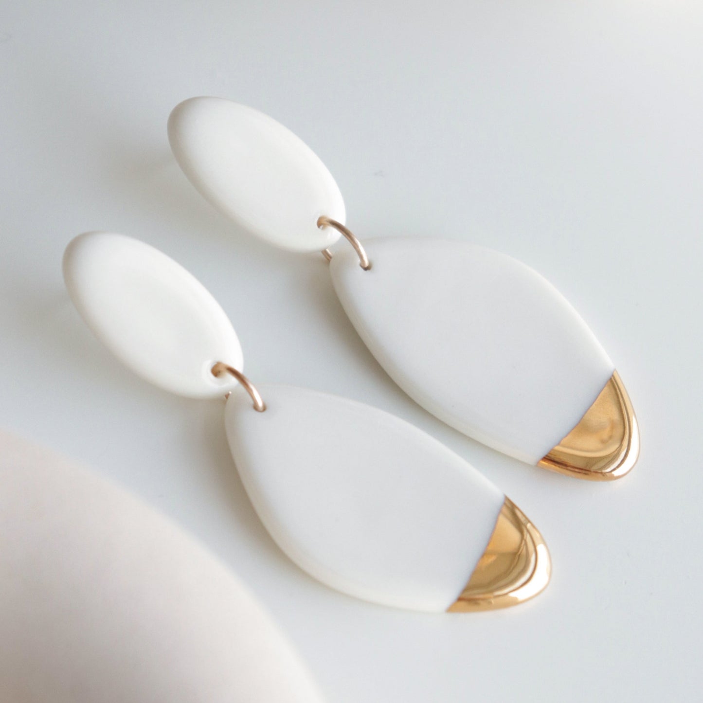 The Flow Earrings by Rozenthal Jewelry are modern, minimalist porcelain statement pieces featuring linked white ovals and a gold-tipped accent, elegantly displayed on a light surface.