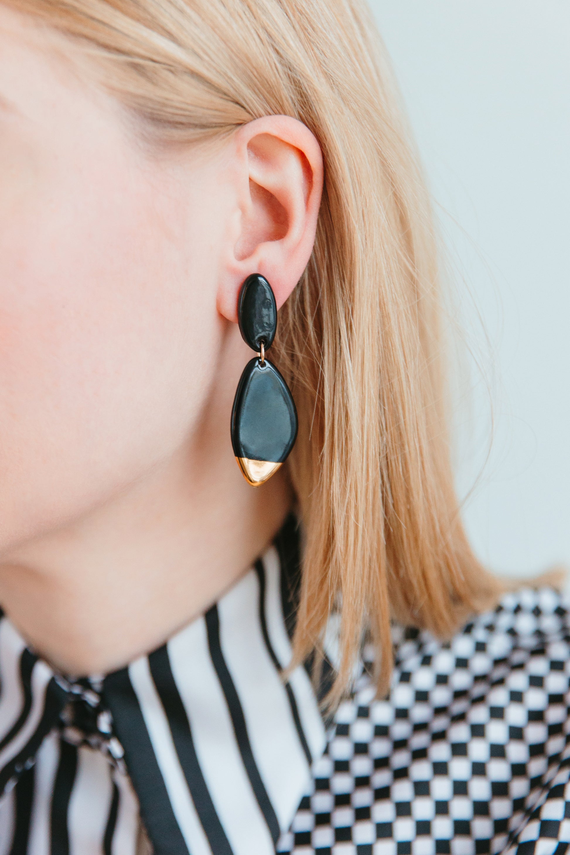 Close-up of a person with blonde hair wearing Rozenthal Jewelry’s Flow Earrings—large, black, teardrop-shaped lightweight waterproof earrings—and a patterned black-and-white shirt with vertical and geometric designs.