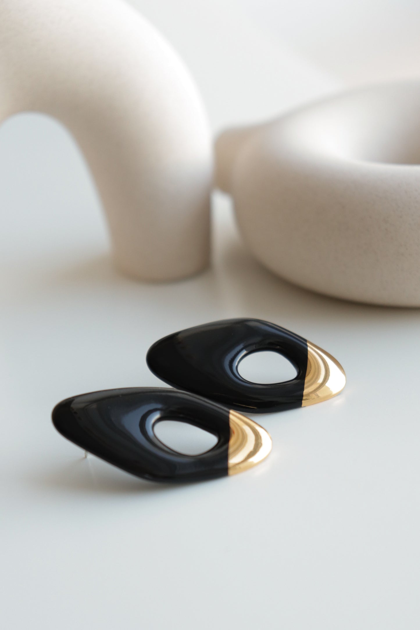 A pair of Flow Earrings by Rozenthal Jewelry—modern, oval-shaped, black and gold, hypoallergenic, and handmade in Latvia—are shown on a white surface with abstract beige ceramic decor in the background.