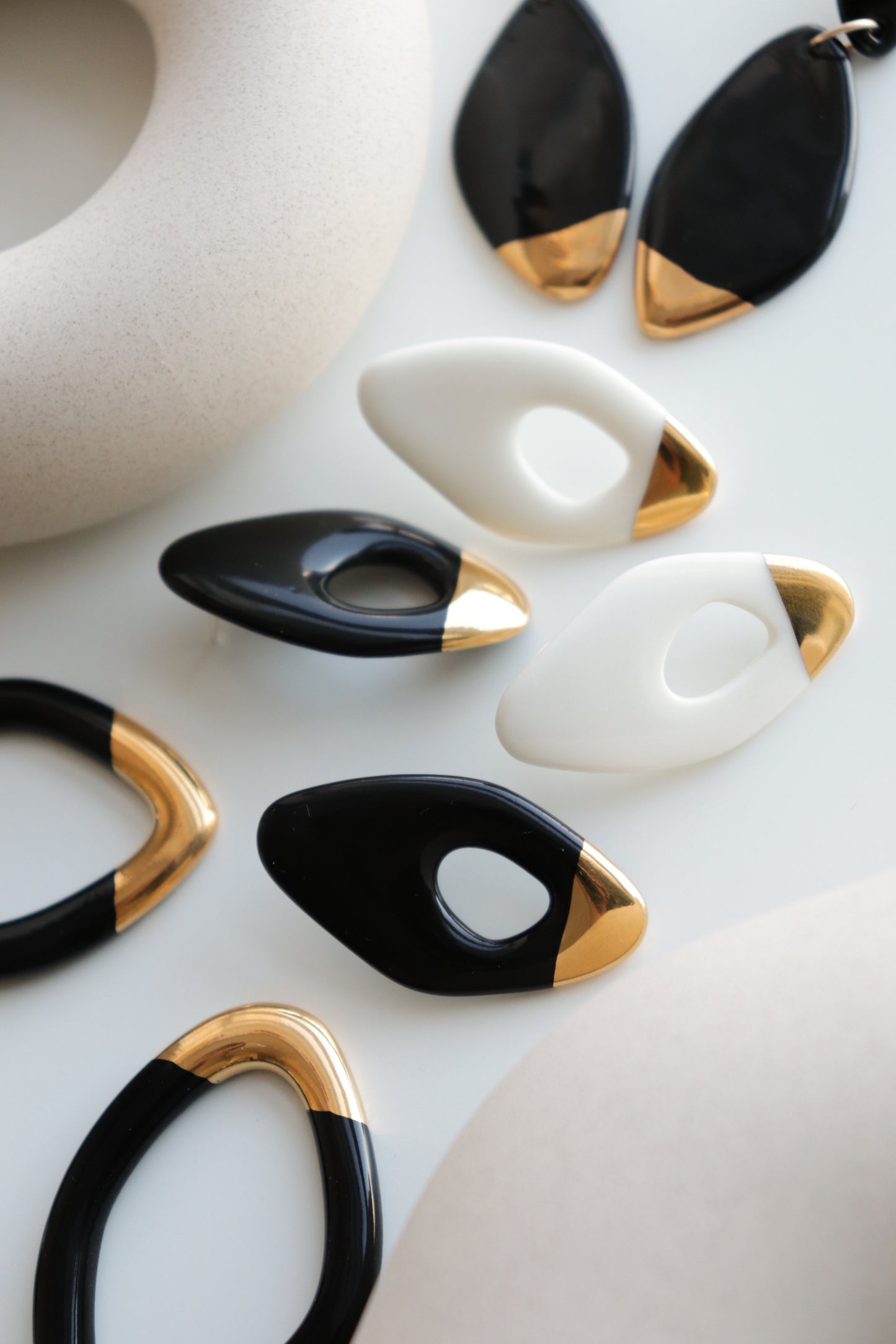 Flow Earrings by Rozenthal Jewelry are modern, oval-shaped hypoallergenic designer earrings in black, white, and gold with glossy finishes and elegant gold accents. Handmade in Latvia, they are artfully arranged on a white surface.