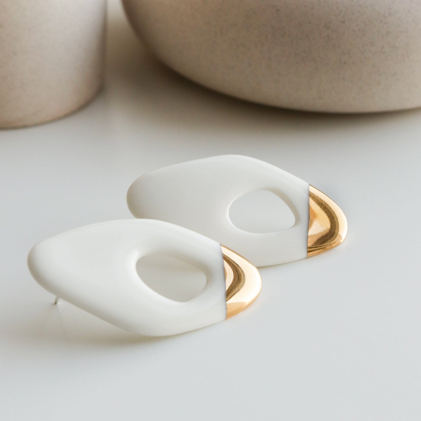 The Flow Earrings by Rozenthal Jewelry, featuring modern white and gold geometric shapes, are displayed on a white surface with soft beige ceramic vases in the background.