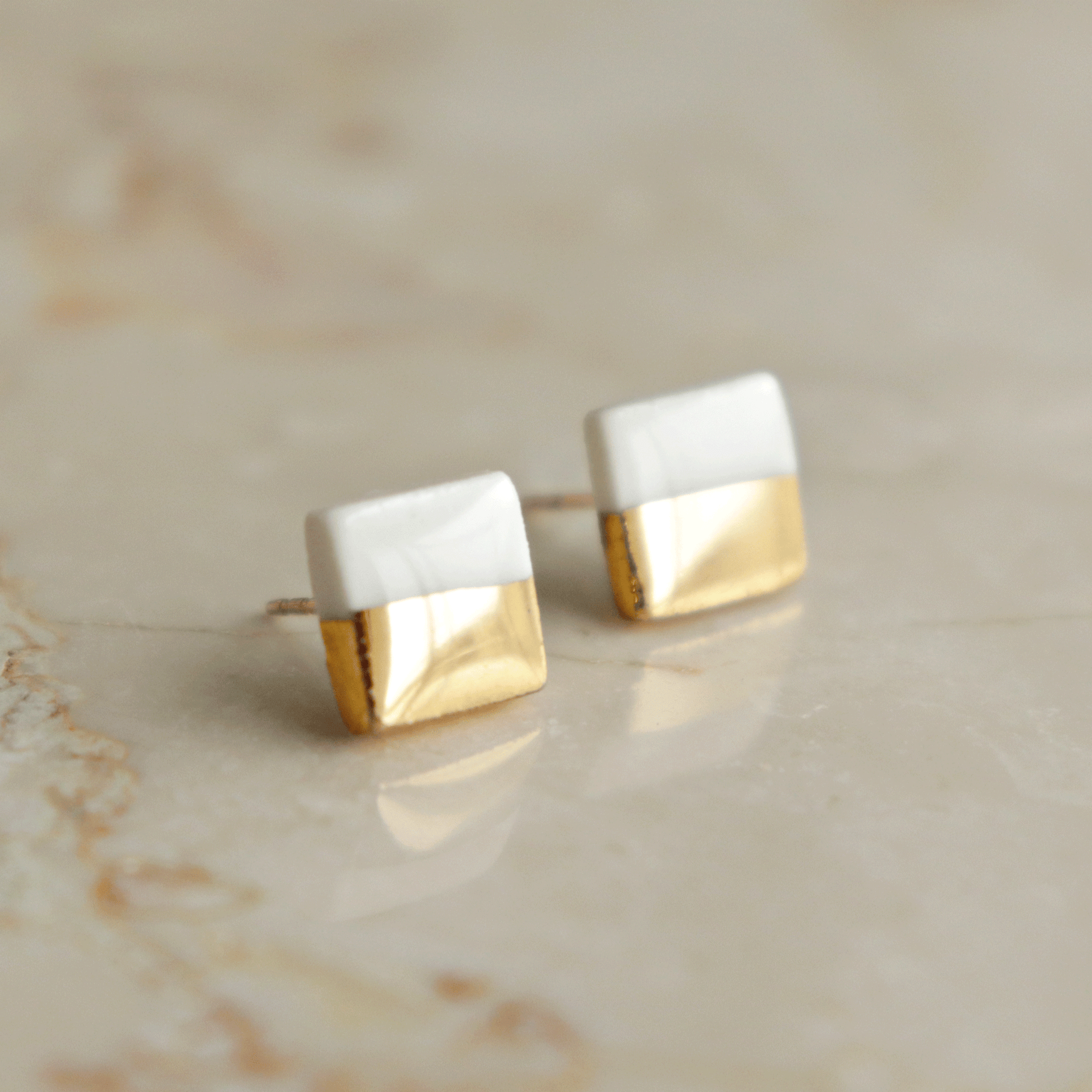 Edgy Studs / XS by Rozenthal Jewelry—square porcelain earrings with a half-white, half-gold design—are handcrafted and displayed on a smooth, light-colored marble surface.