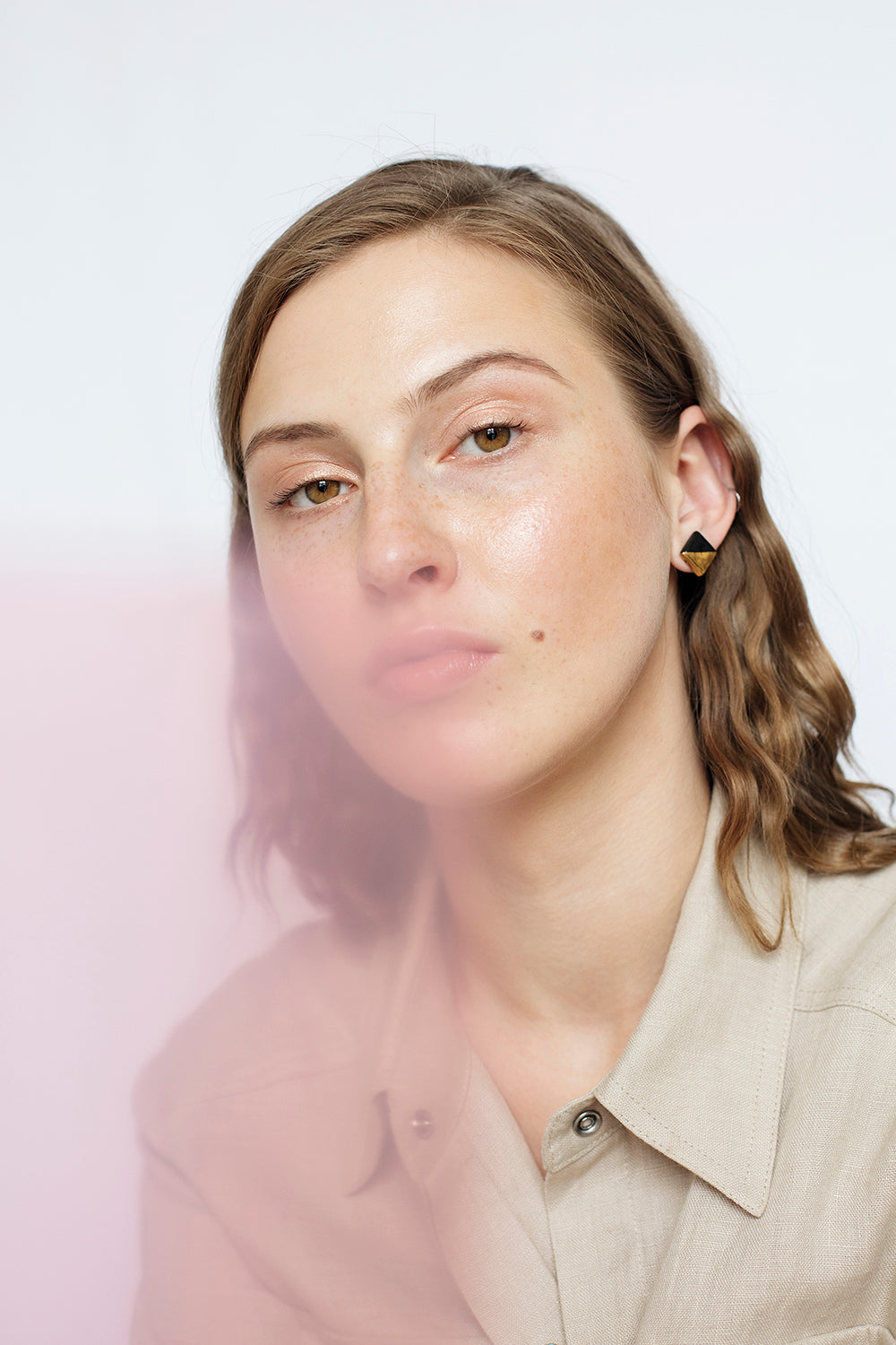 A woman with wavy light brown hair and a beige shirt looks at the camera, wearing Rozenthal Jewelrys Edgy Studs / S—minimalist geometric gold earrings. A soft pink blur appears in the foreground against a light background.