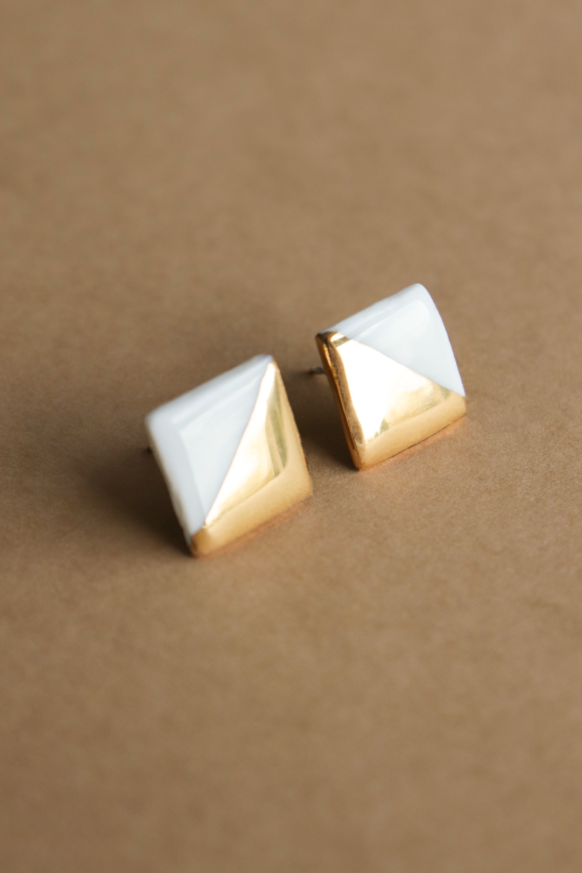 Edgy Studs / S by Rozenthal Jewelry—square stud earrings with a geometric design, featuring a diagonal split of shiny gold and glossy white, shown on a light brown surface. Perfect minimalism for jewelry lovers.