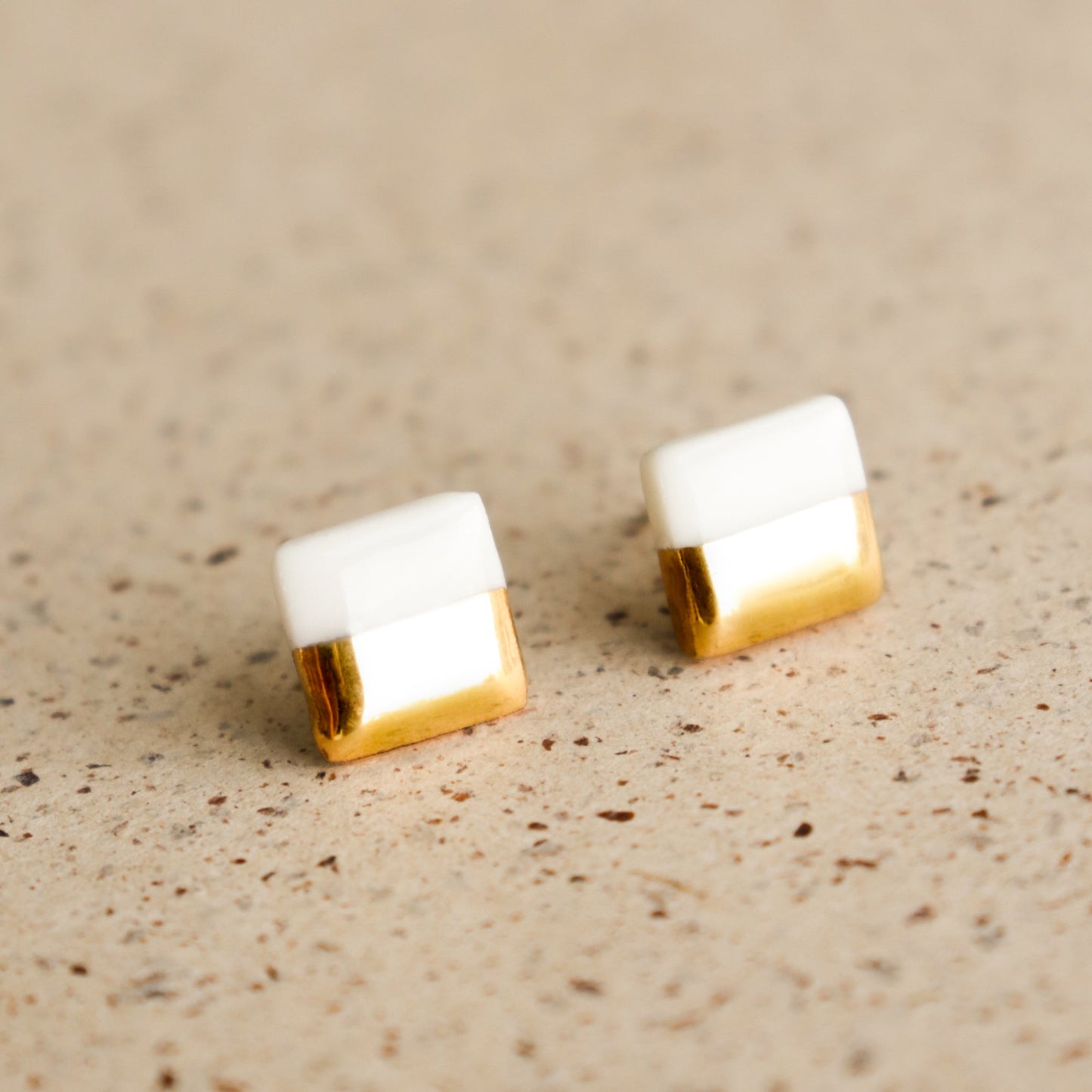 The Edgy Studs / XS by Rozenthal Jewelry are minimalist, small square earrings featuring a white top half and shiny gold bottom half, displayed elegantly on a speckled beige surface.