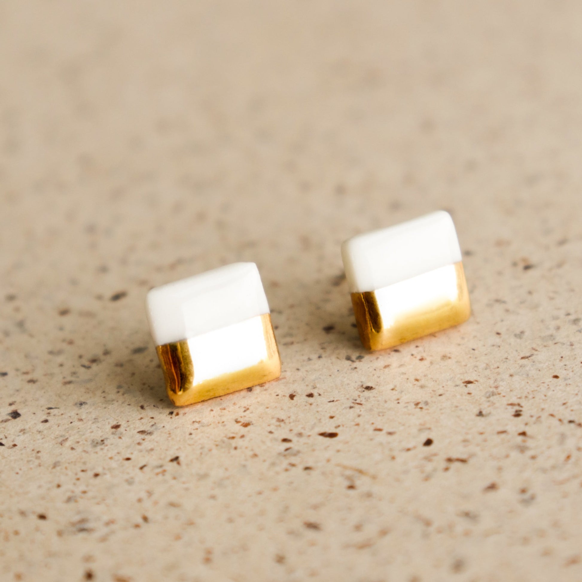 The Edgy Studs / XS by Rozenthal Jewelry are minimalist, small square earrings featuring a white top half and shiny gold bottom half, displayed elegantly on a speckled beige surface.