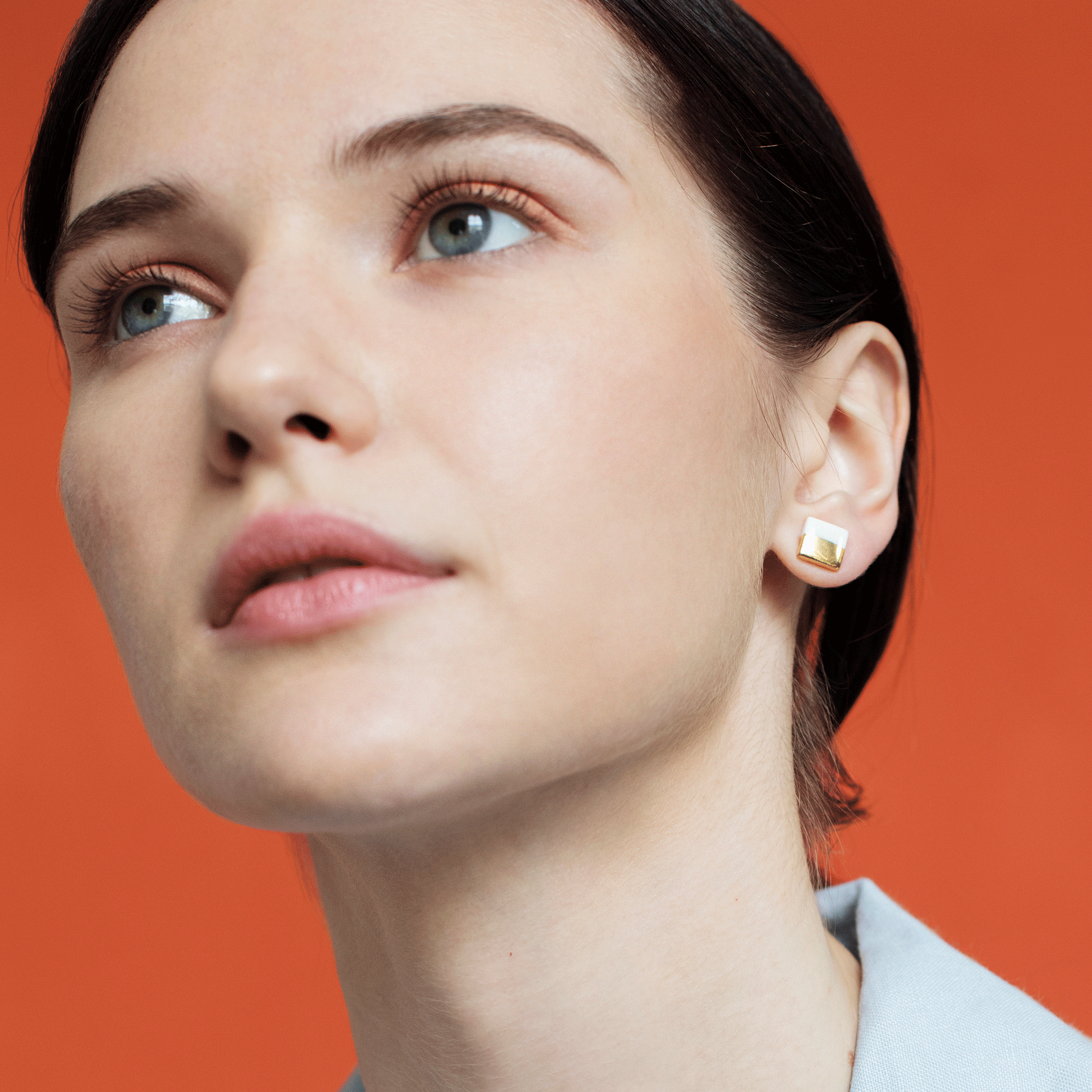 A fair-skinned woman with blue eyes and dark hair pulled back, wearing minimal makeup and a light blue shirt, looks upward. She showcases Rozenthal Jewelry’s Edgy Studs / XS—hypoallergenic square gold designer earrings—against an orange background.