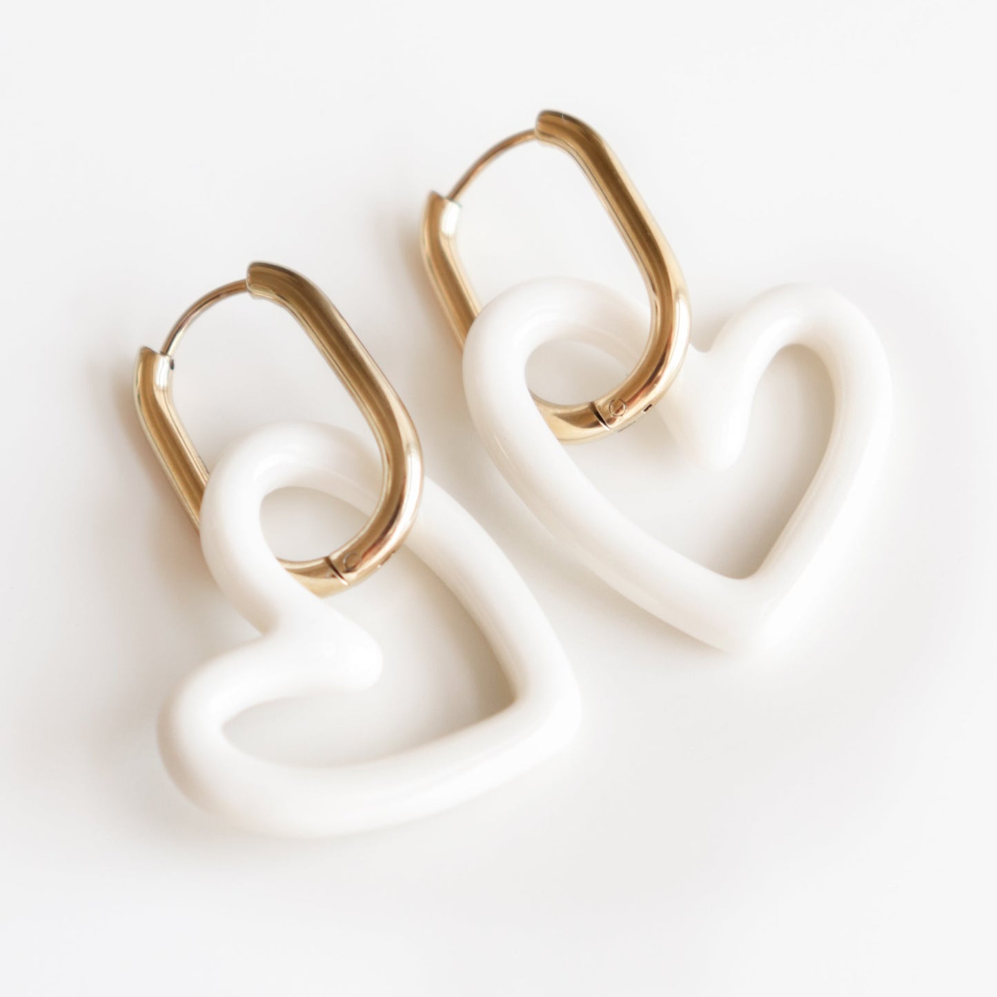 The Heart Earrings by Rozenthal Jewelry are handmade gold hoops with white, heart-shaped porcelain pendants, displayed on a white background. These statement earrings offer a unique touch to any outfit.