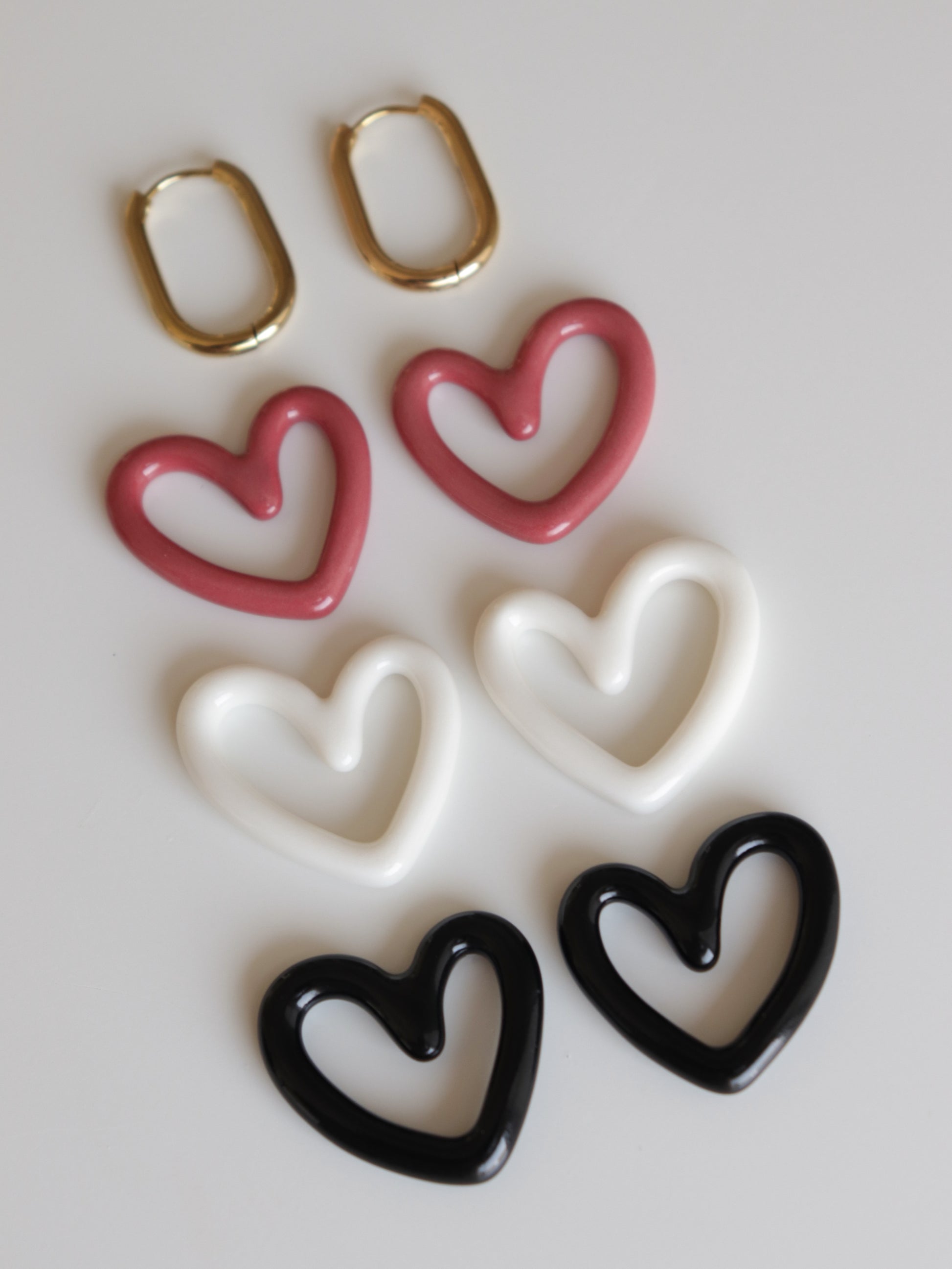 A pair of gold oval Heart Earrings by Rozenthal Jewelry with lightweight, waterproof hoops and six heart-shaped charms in pink, white, and black, arranged in pairs on a white surface.