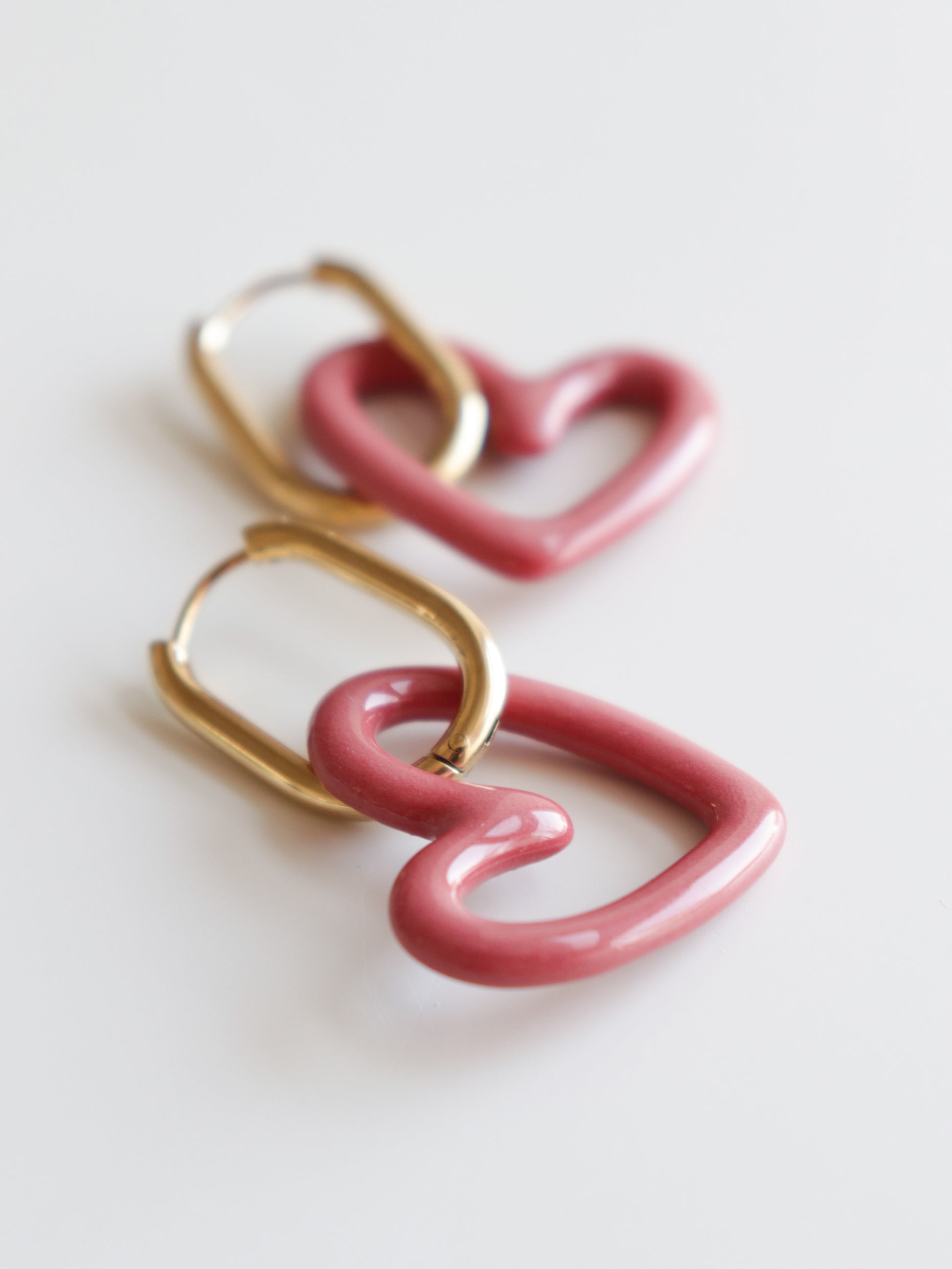 The Heart Earrings by Rozenthal Jewelry are gold hoops with glossy pink heart-shaped pendants. These hypoallergenic designer earrings are shown on a white surface.