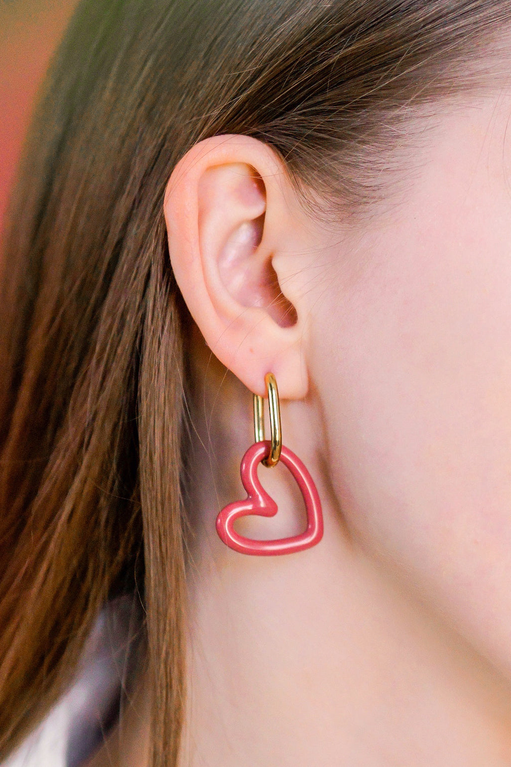 Close-up of a person wearing Rozenthal Jewelrys Heart Earrings—gold hoops with pink, heart-shaped pendants—partially hidden by brown hair. These hypoallergenic designer earrings are perfect for sensitive ears and add a touch of elegance.