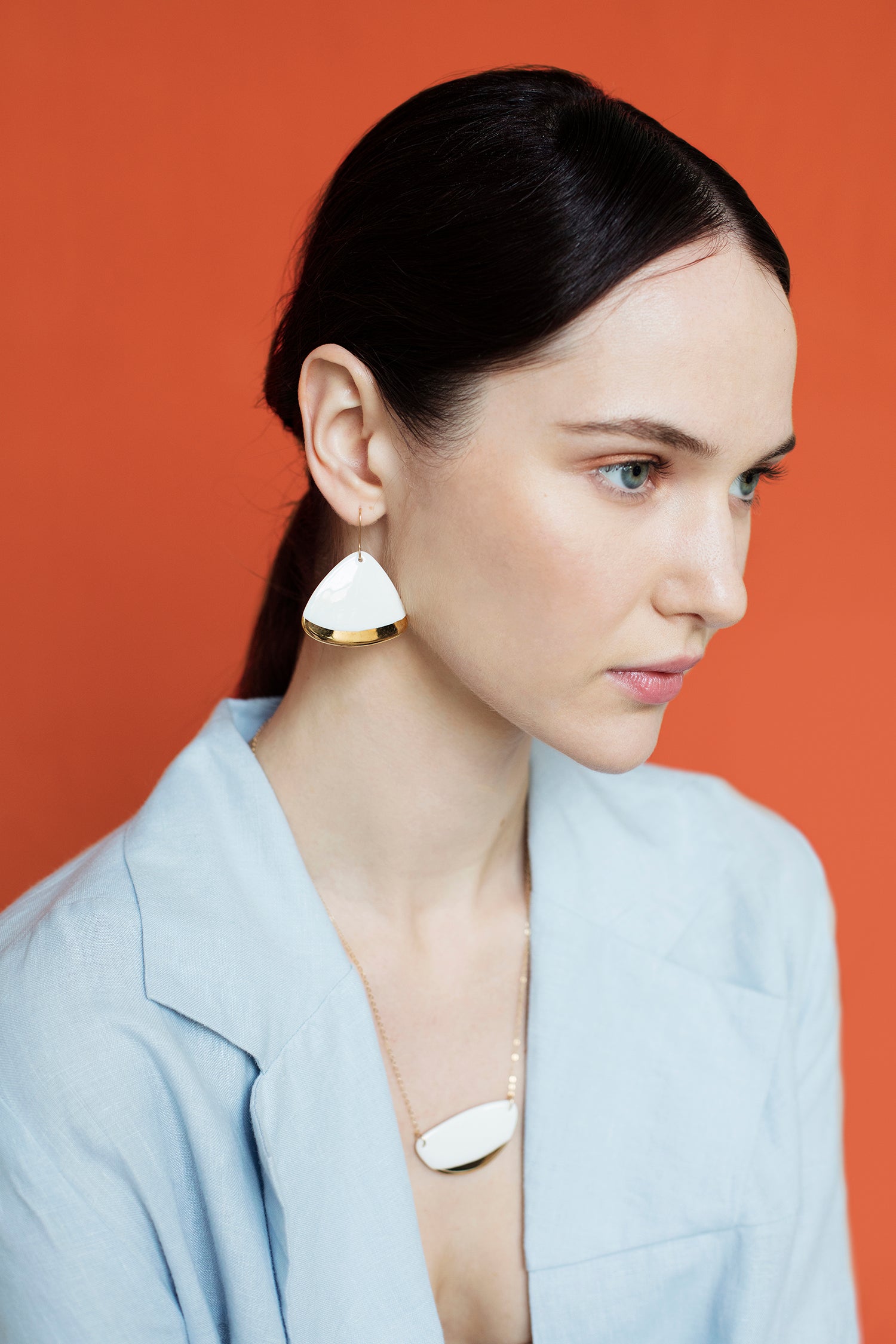 A woman with fair skin and dark hair pulled back wears a light blue blazer and Rozenthal Jewelry’s Leaf Earrings—striking white and gold statement pieces—posing against an orange background.