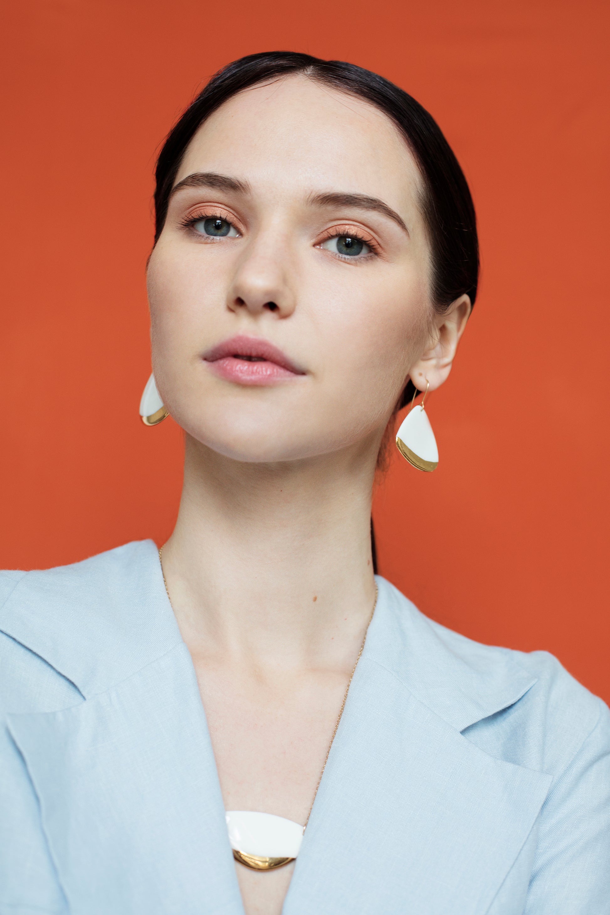 A woman with dark hair pulled back wears a light blue blazer and poses against an orange background, accessorized with Rozenthal Jewelry’s Leaf Earrings in white and gold geometric design, along with a matching necklace.