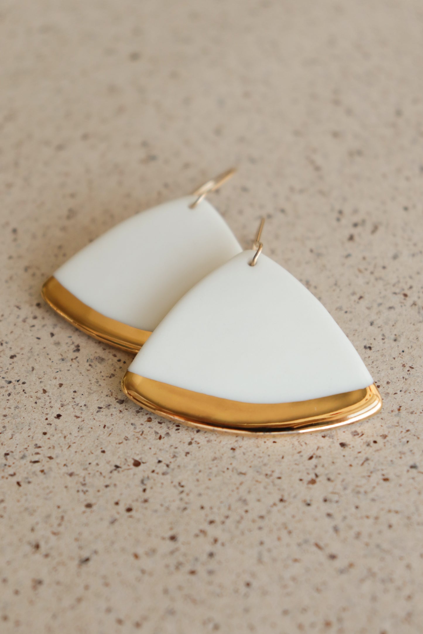 Leaf Earrings by Rozenthal Jewelry feature a smooth white surface with shiny gold edges in a minimalist triangular design, perfect for modern jewelry lovers, displayed against a speckled beige background.