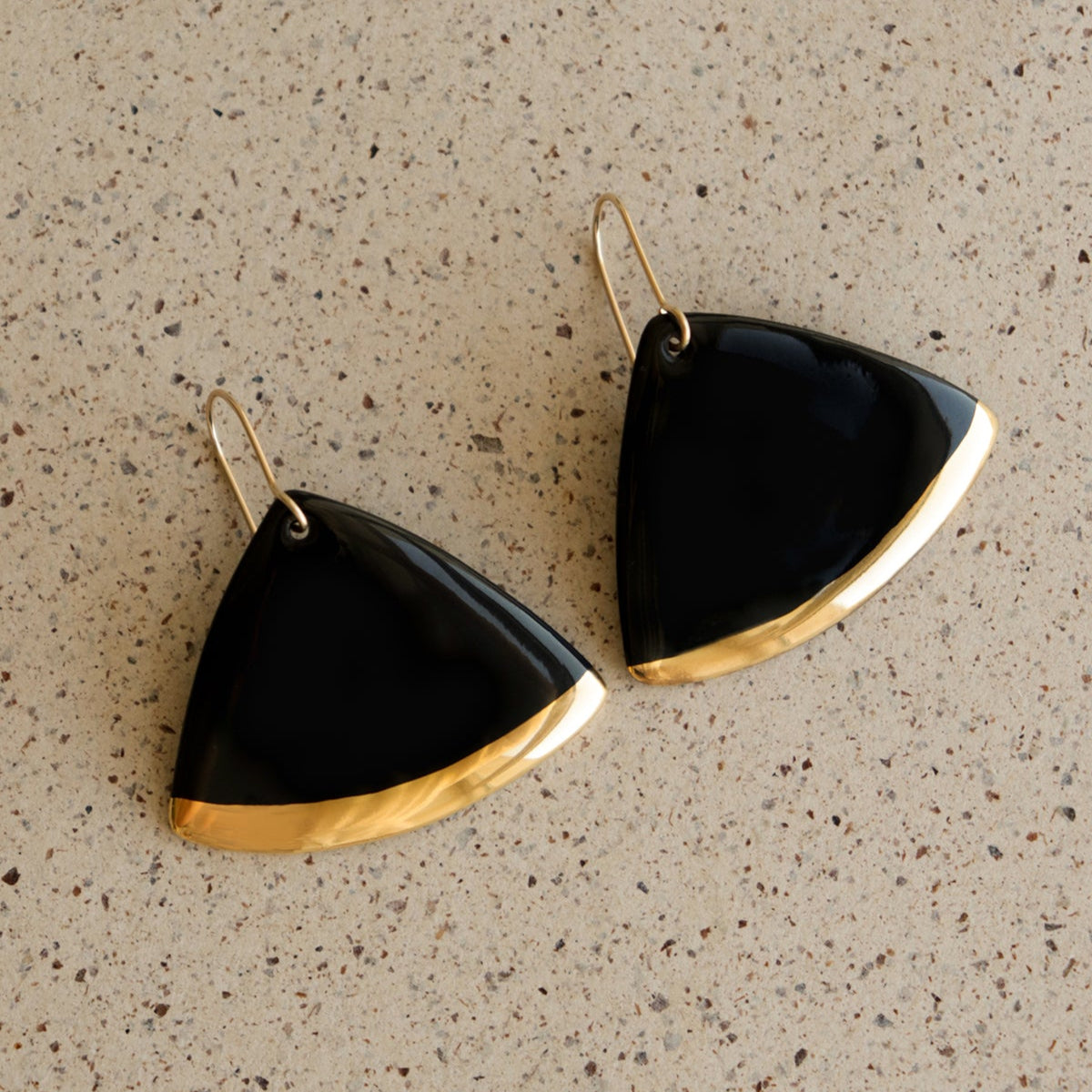The Leaf Earrings by Rozenthal Jewelry feature black, triangular pendants with gold trim along the bottom edge, displayed on a speckled beige surface—an elegant showcase of minimalist design.
