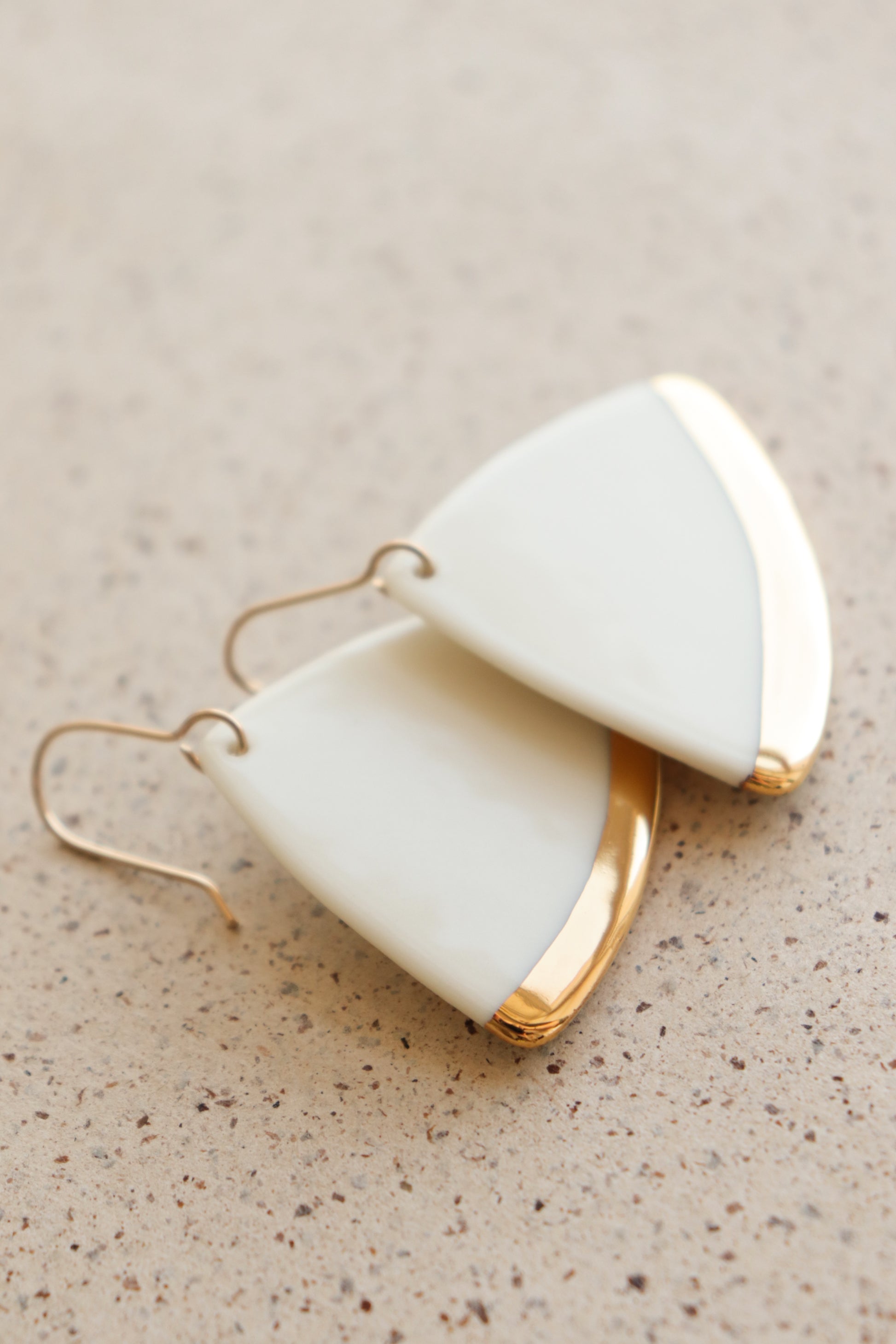Leaf Earrings by Rozenthal Jewelry: These minimalist white porcelain earrings feature a triangular leaf design with gold accents and hooks, displayed on a speckled beige surface.