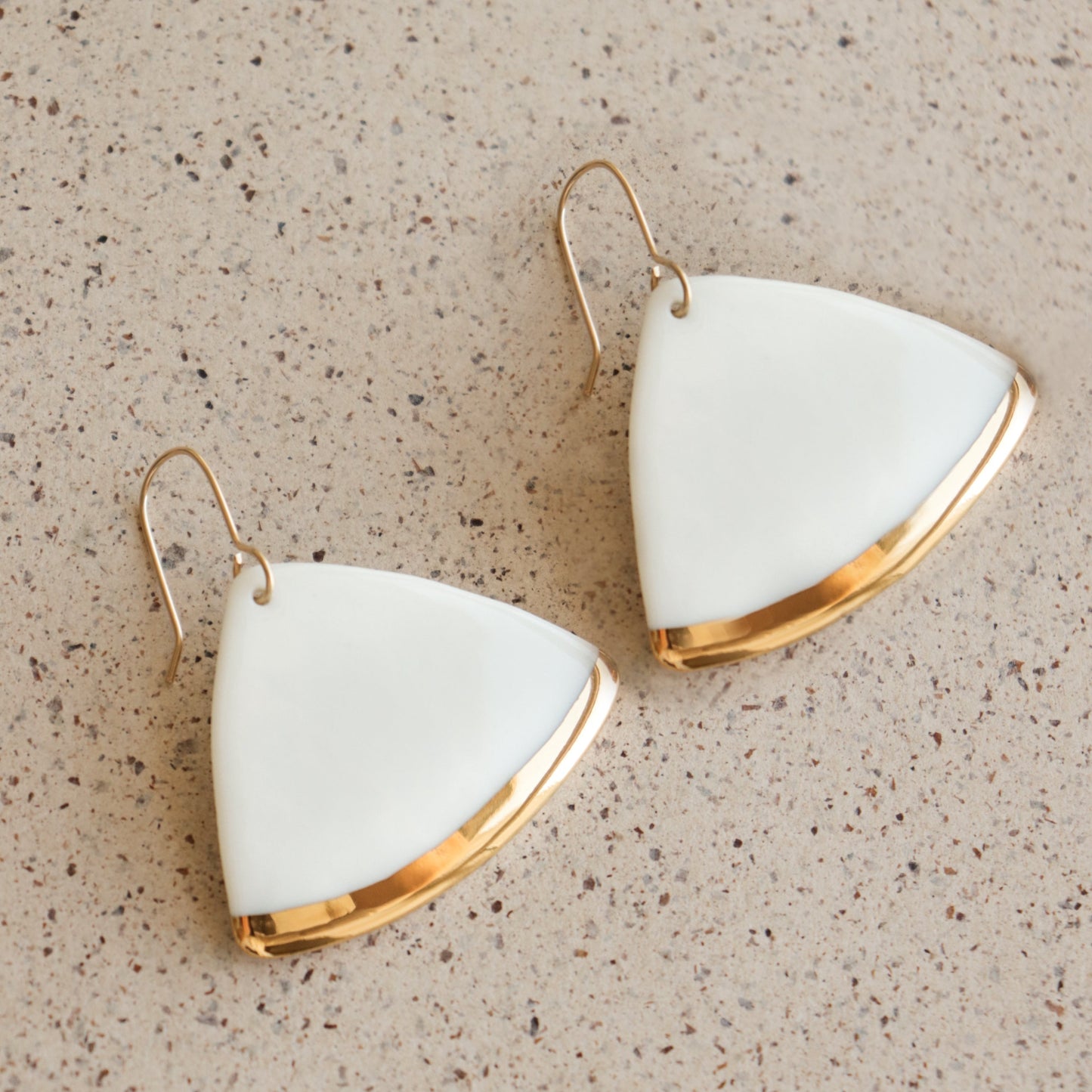 The Leaf Earrings by Rozenthal Jewelry are handmade, minimalist white earrings with a triangular shape, gold edges, and hooks, displayed on a speckled beige surface.