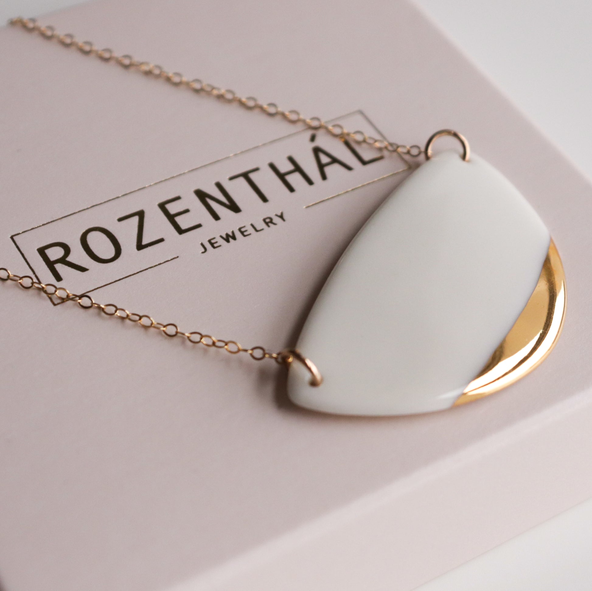 The Rozenthal Jewelry Leaf Necklace, featuring a minimalist gold chain with a geometric porcelain pendant, is elegantly displayed on a pale pink jewelry box.