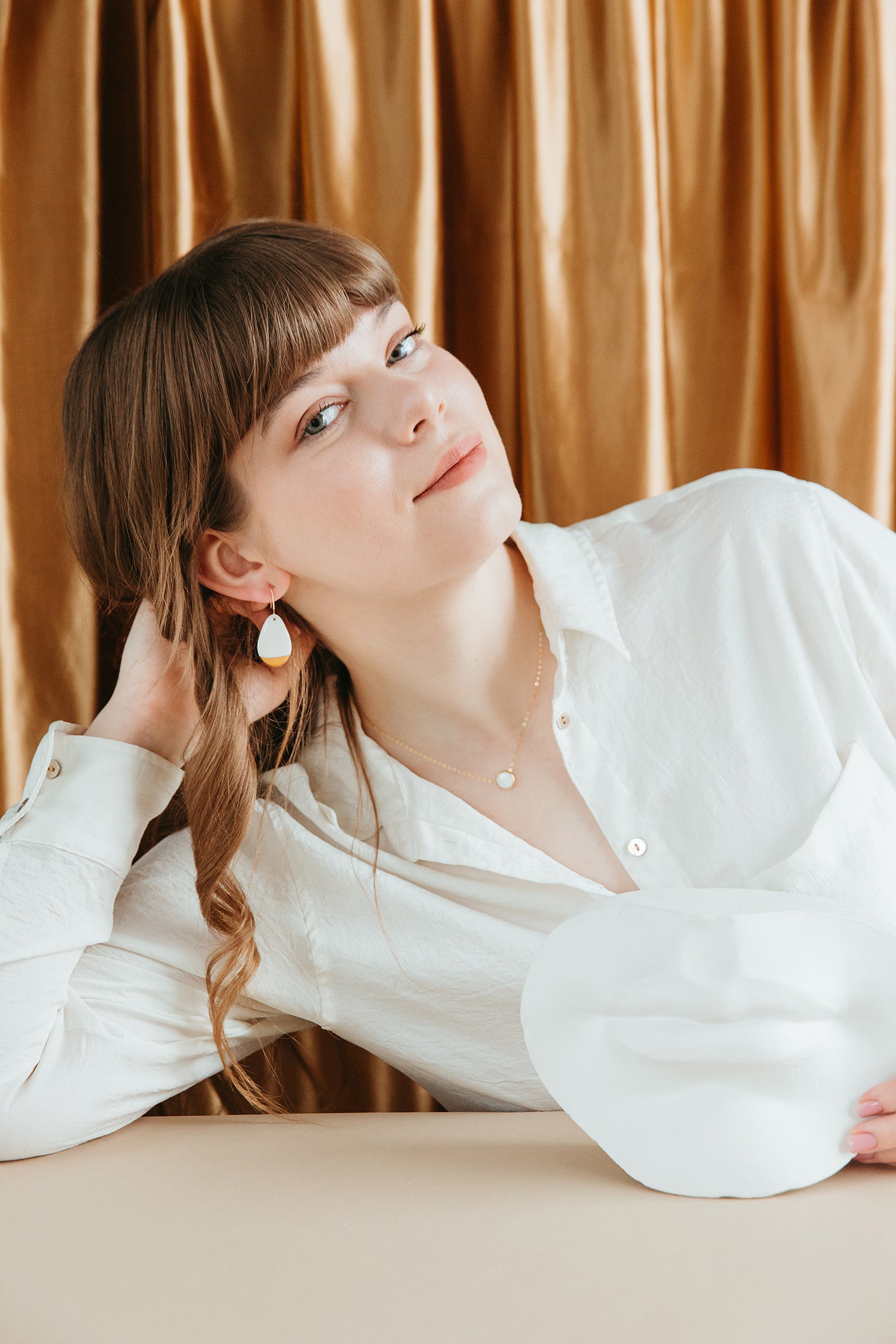A woman with light brown hair and bangs, wearing a white shirt and the Rozenthal Jewelry Round Necklace, leans on a table with one hand in her hair. A white facial mask is on the table, and gold curtains hang in the background.