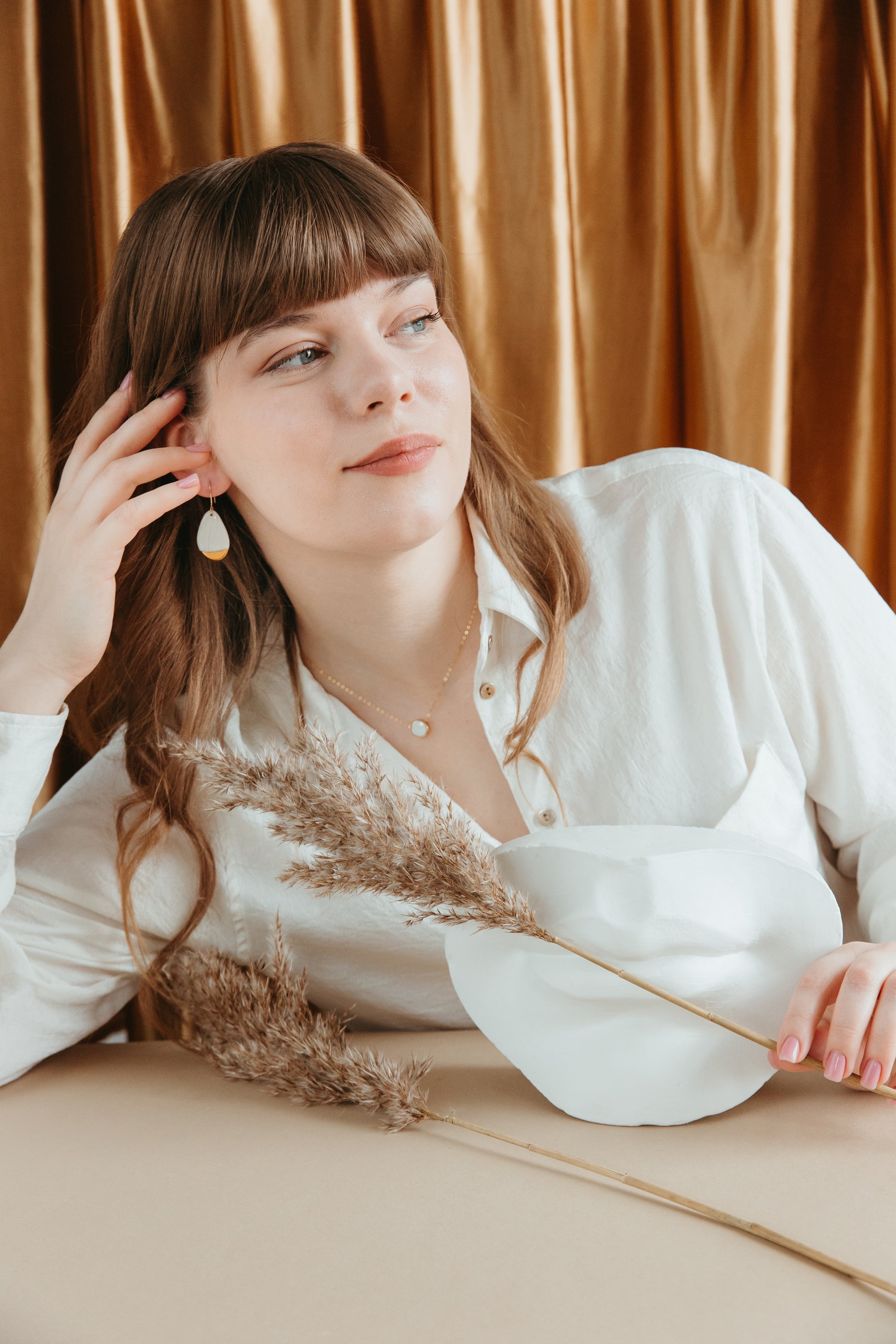 A woman with light brown hair in a white blouse sits at a beige table decorated with dried grass and a white mask, wearing the Rozenthal Jewelry Round Necklace, gazing thoughtfully to the side before golden curtains.
