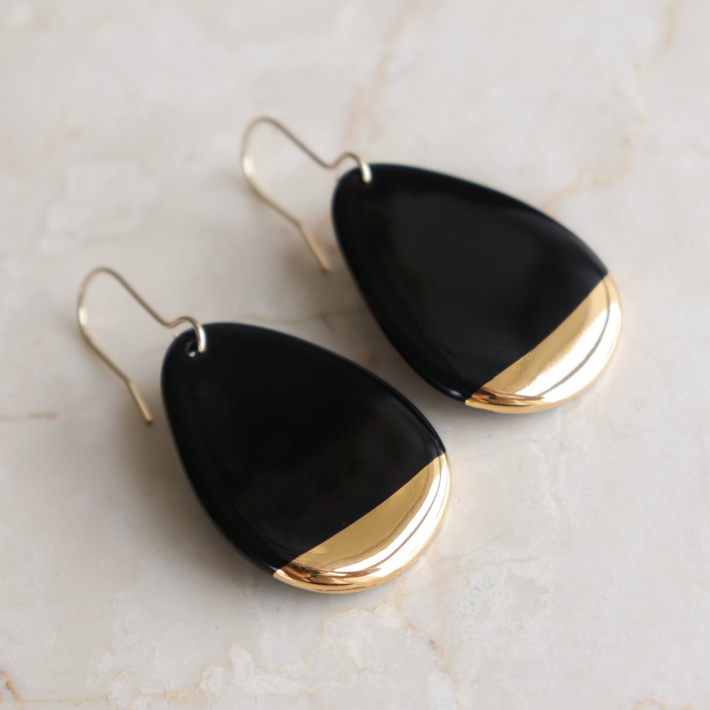Rozenthal Jewelry’s Drop Earrings / M are teardrop-shaped porcelain statement earrings with black and gold accents, gold hooks, and a glossy finish. Handmade in Latvia, they’re lightweight, waterproof, and shown on a light marble surface.