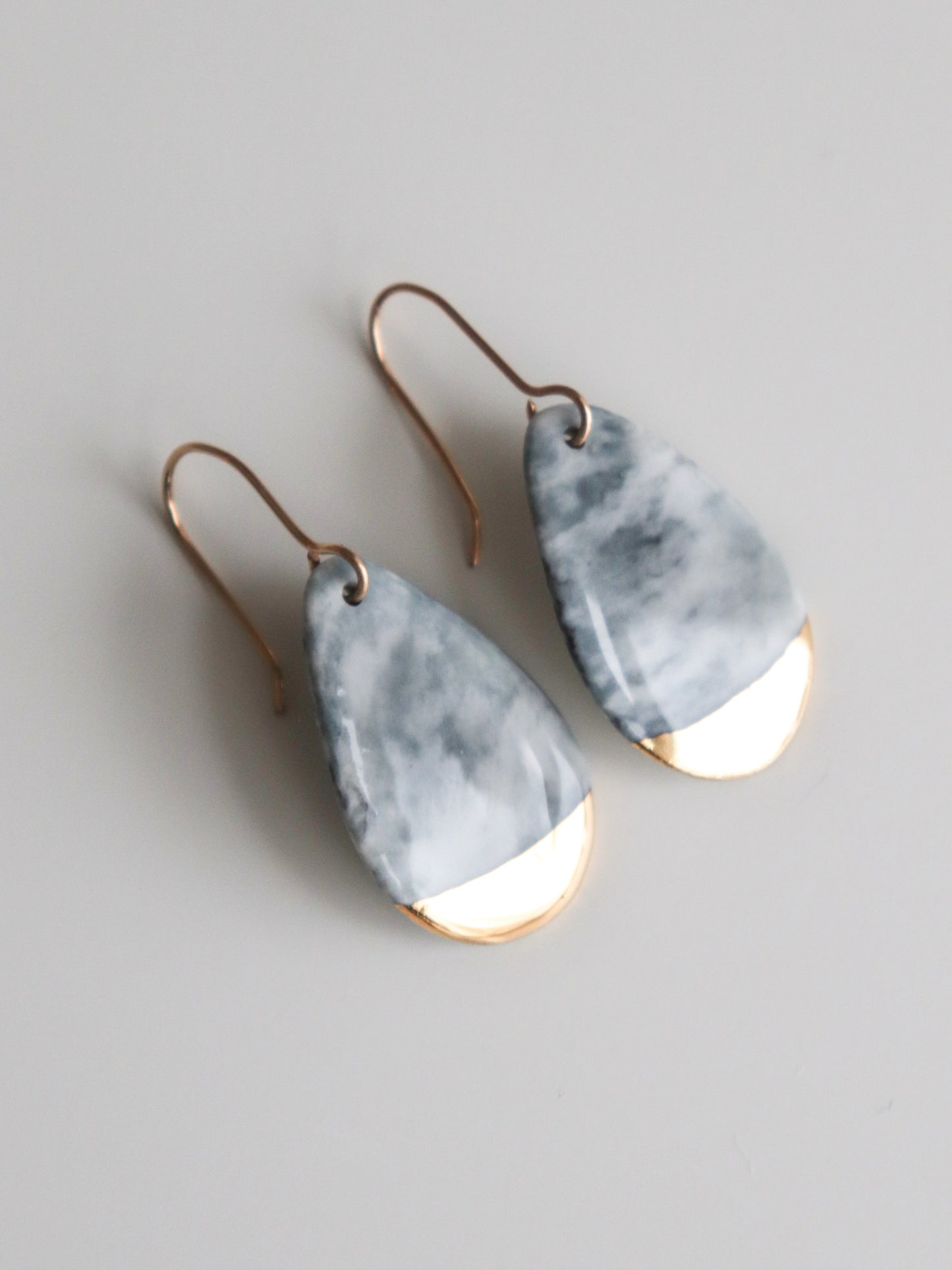 The Drop Earrings / S by Rozenthal Jewelry are handmade in Latvia, featuring marbled gray and white teardrop tops with gold-toned metal bottoms, shown resting on a light surface.