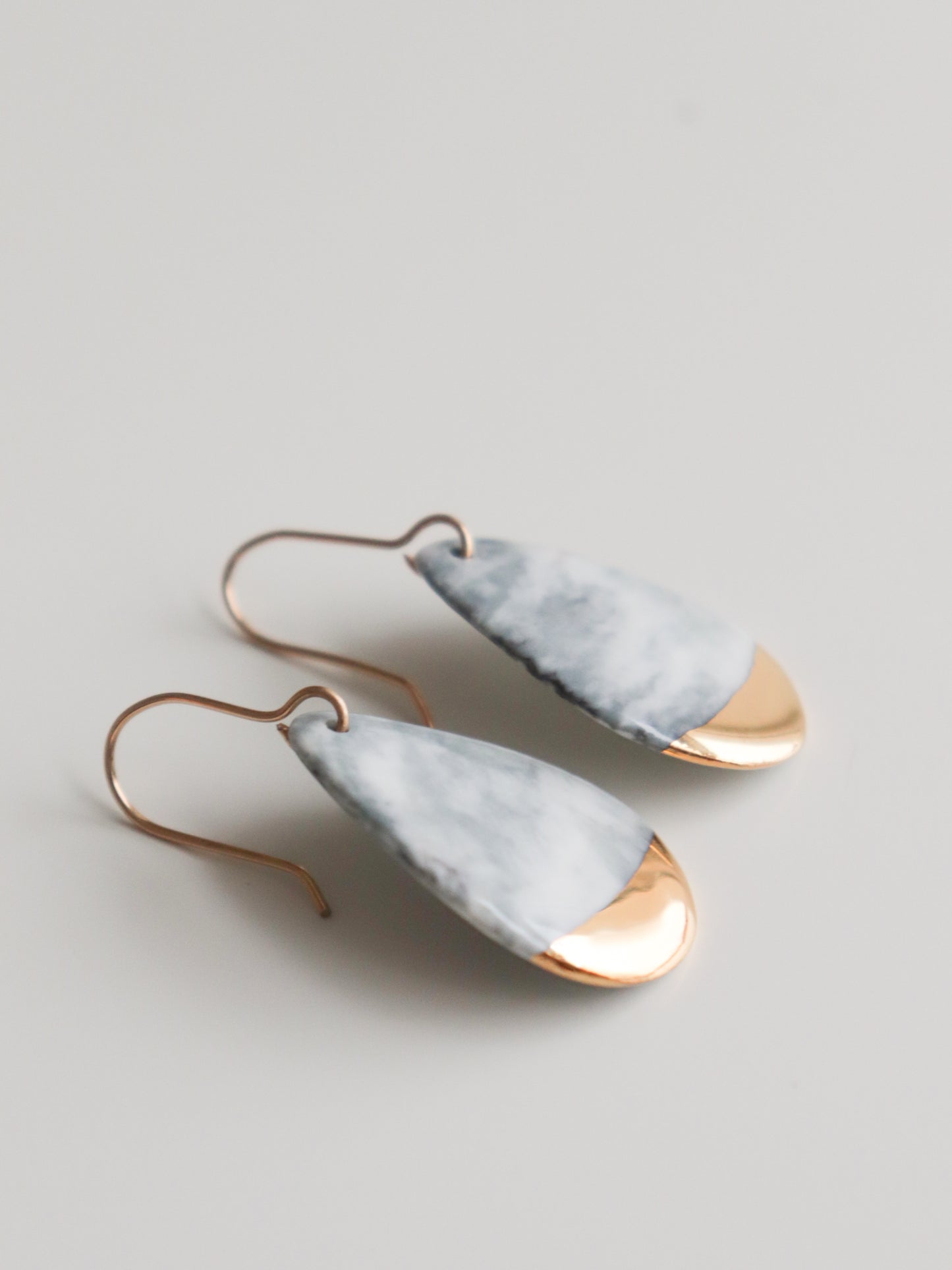 The Drop Earrings / S by Rozenthal Jewelry are handmade in Latvia with gold hooks, featuring teardrop shapes with marbled white and gray tops and shiny gold bottoms. Displayed on a plain white background.