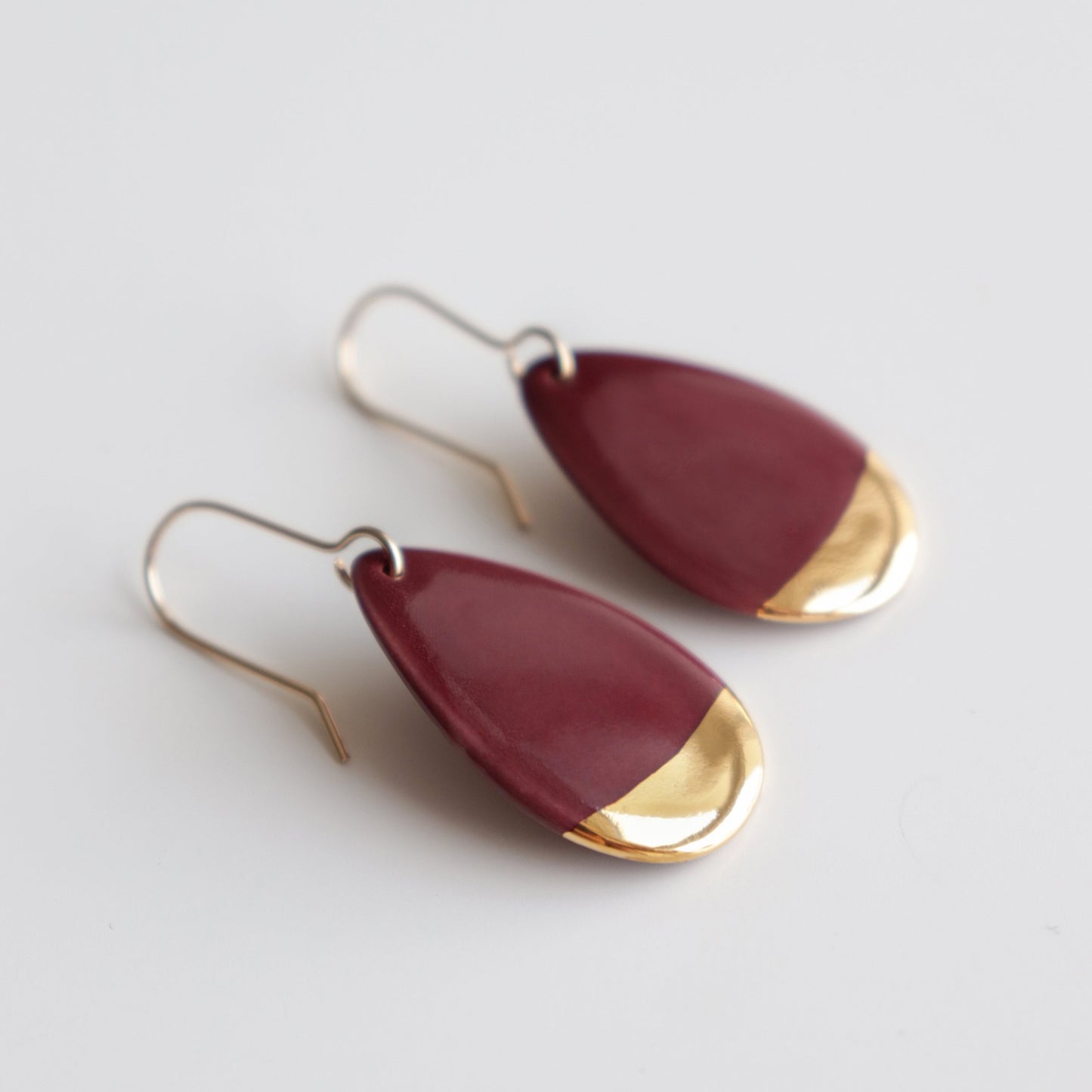 Rozenthal Jewelry’s Drop Earrings / S are teardrop-shaped handmade porcelain earrings featuring a maroon body with gold accents at the bottom, displayed on a white surface.