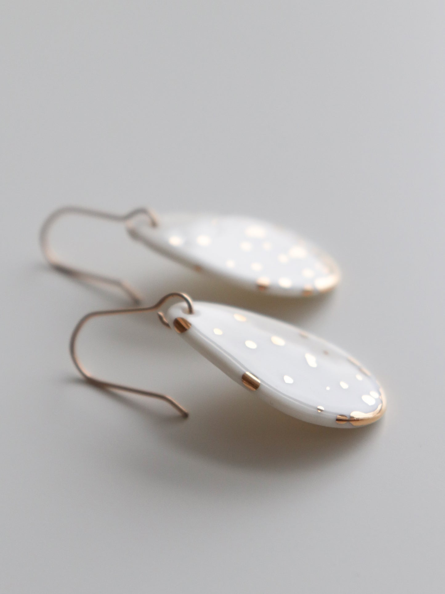The Rozenthal Jewelry Drop Earrings / S are white, oval-shaped handmade earrings with small gold dots and gold edges, featuring simple gold hooks. Hypoallergenic designer pieces crafted in Latvia rest on a smooth, light gray surface.
