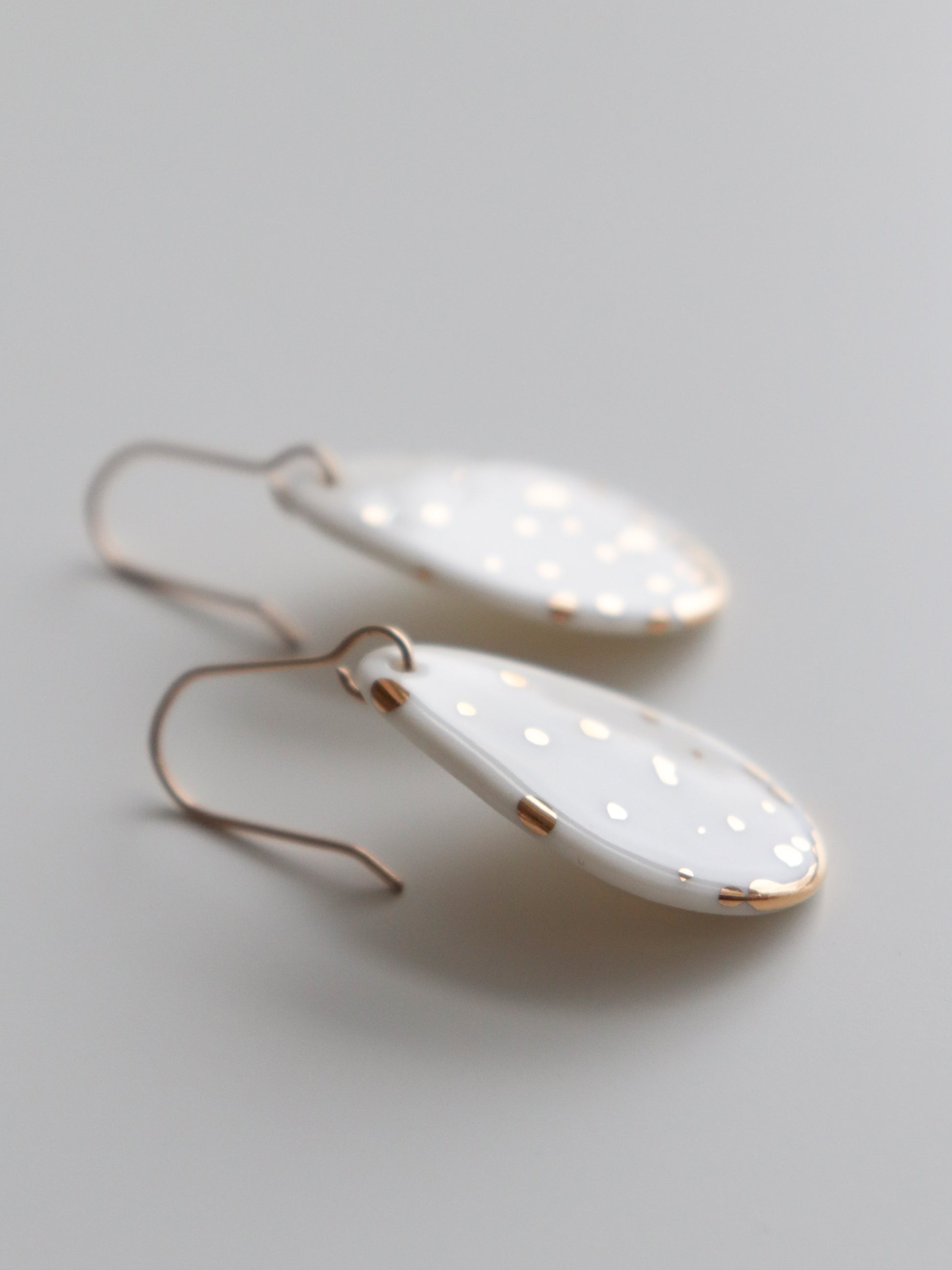 The Rozenthal Jewelry Drop Earrings / S are white, oval-shaped handmade earrings with small gold dots and gold edges, featuring simple gold hooks. Hypoallergenic designer pieces crafted in Latvia rest on a smooth, light gray surface.