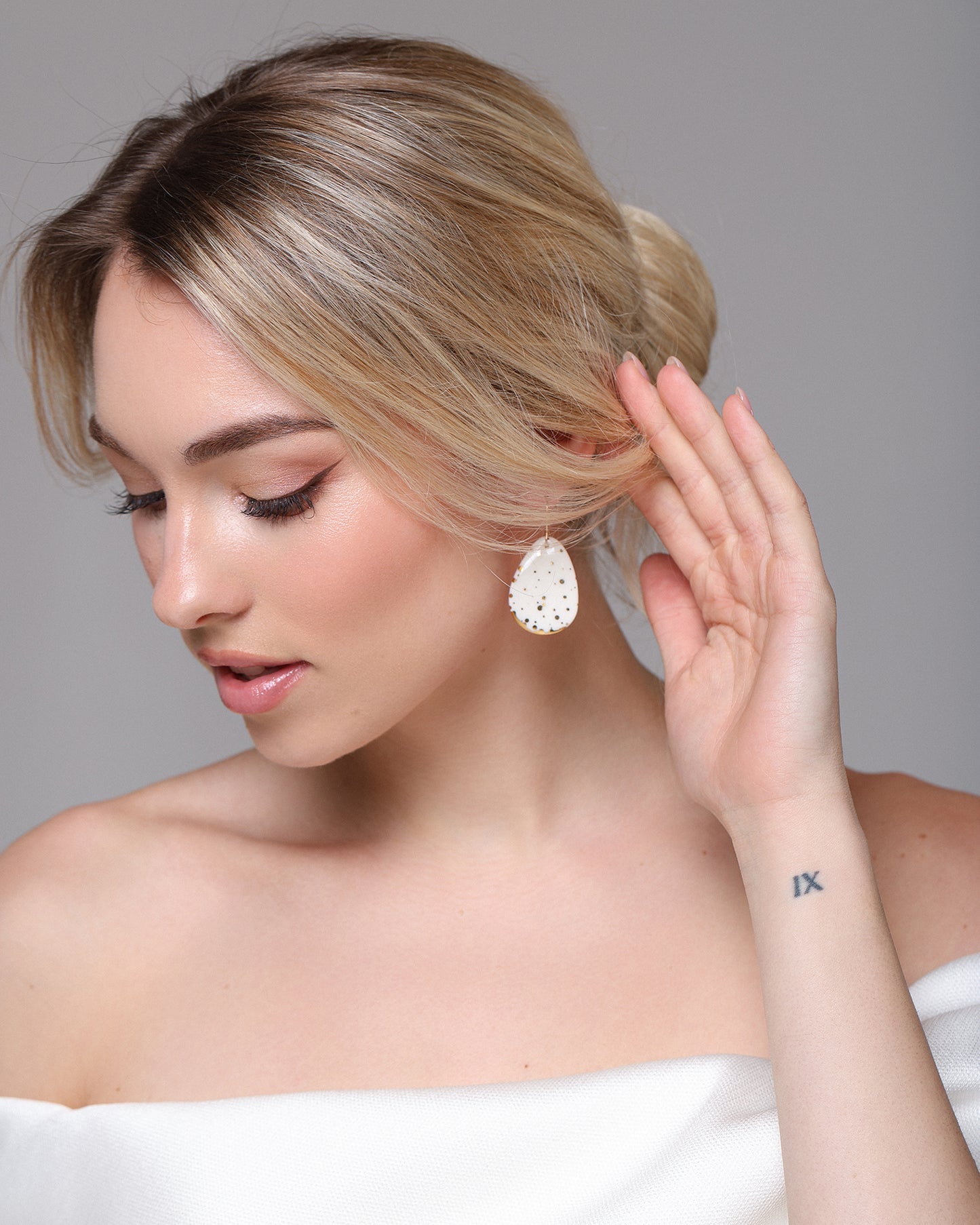 A blonde woman in a low bun, wearing a white off-shoulder top and natural makeup, touches her ear adorned with Rozenthal Jewelrys handmade Drop Earrings / M; a Roman numeral IX tattoo is visible on her wrist.