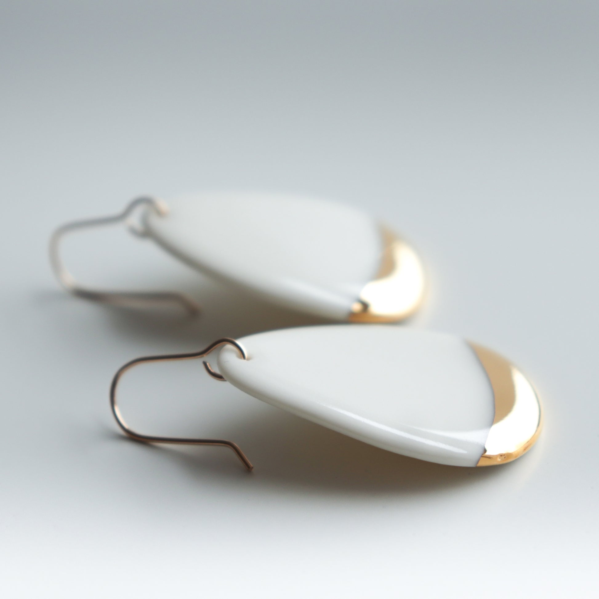 Drop Earrings / M by Rozenthal Jewelry: handmade teardrop-shaped porcelain earrings with a white surface, gold-tipped accents, and simple metal hooks. Crafted in Latvia and shown on a light background.