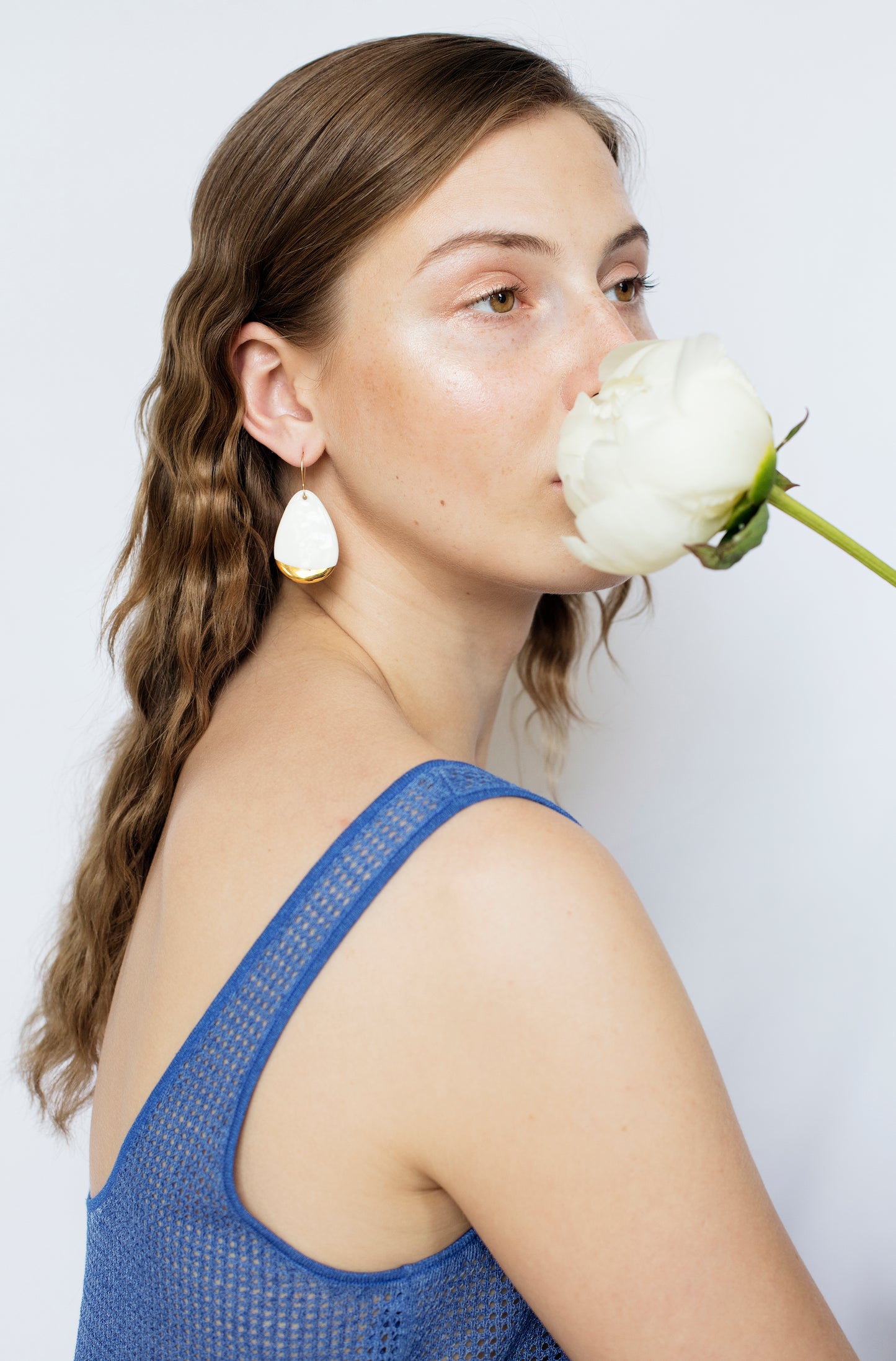 A woman with wavy brown hair wears a blue sleeveless top and Rozenthal Jewelrys Drop Earrings / M, holding a white flower near her face as she gazes off to the side against a plain white background.