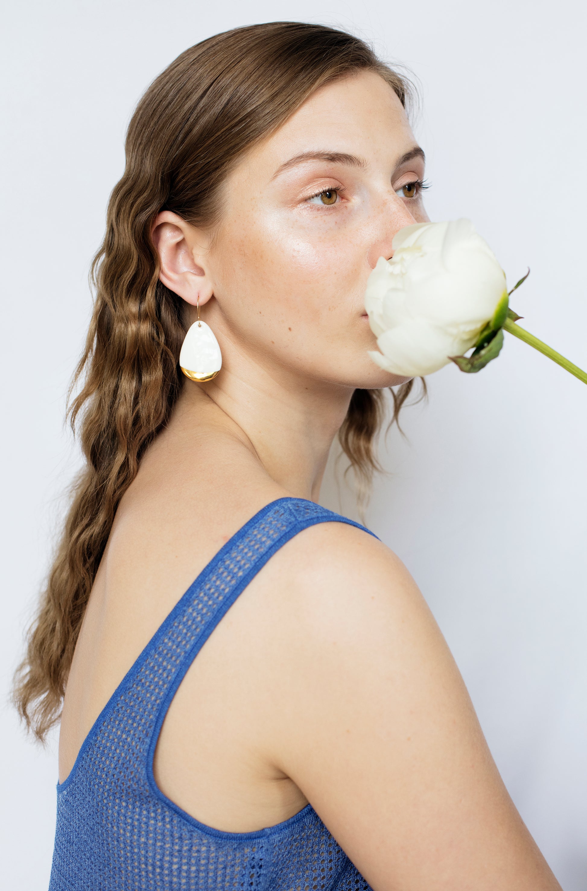 A woman with wavy brown hair wears a blue sleeveless top and Rozenthal Jewelrys Drop Earrings / M, holding a white flower near her face as she gazes off to the side against a plain white background.