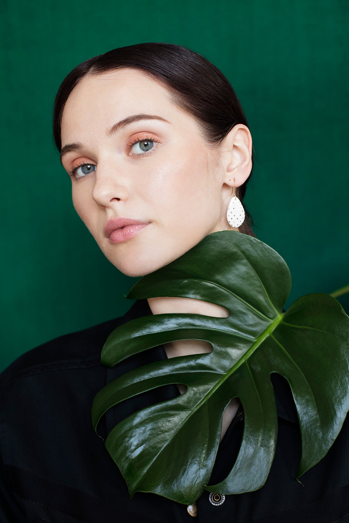 A fair-skinned, dark-haired woman poses before a green background, wearing a black top and eye-catching white Drop Earrings / S by Rozenthal Jewelry—handmade in Latvia—while holding a large monstera leaf under her chin.