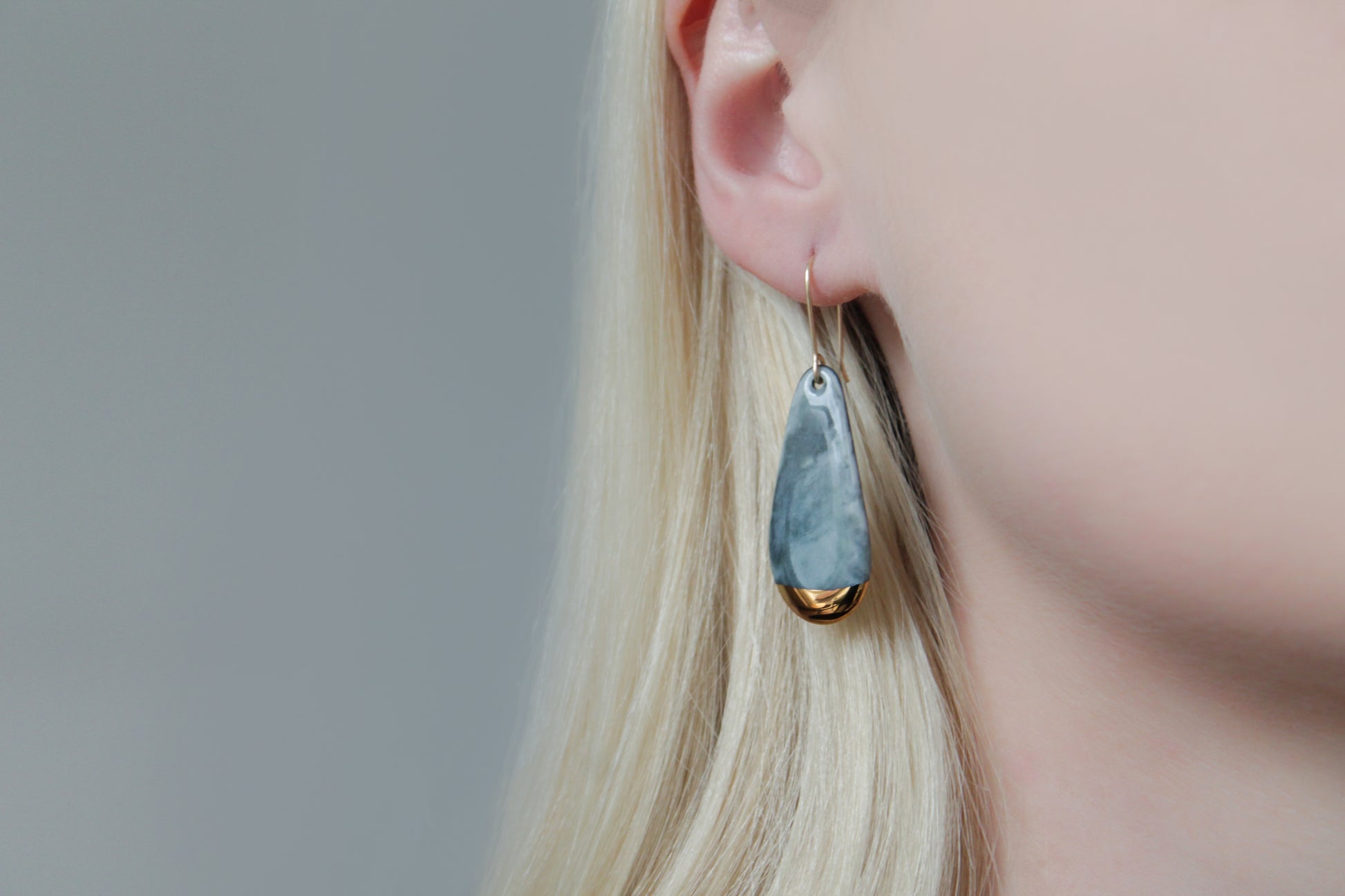 Close-up of a person with light blonde hair wearing Rozenthal Jewelry’s Drop earrings / S—teardrop-shaped statement earrings in marbled gray and gold—set against a neutral gray background.