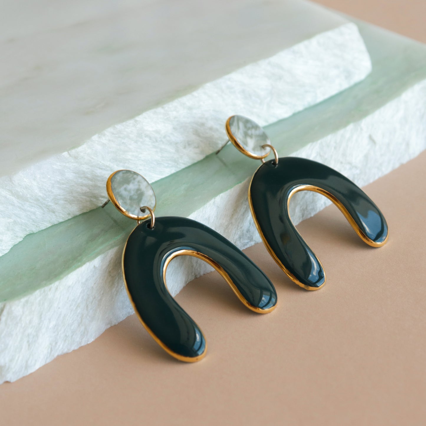 The Arquus Earrings by Rozenthal Jewelry feature marbled porcelain oval studs and glossy dark green U-shaped dangles edged in 24k gold, shown on layered stone slabs with a beige backdrop.
