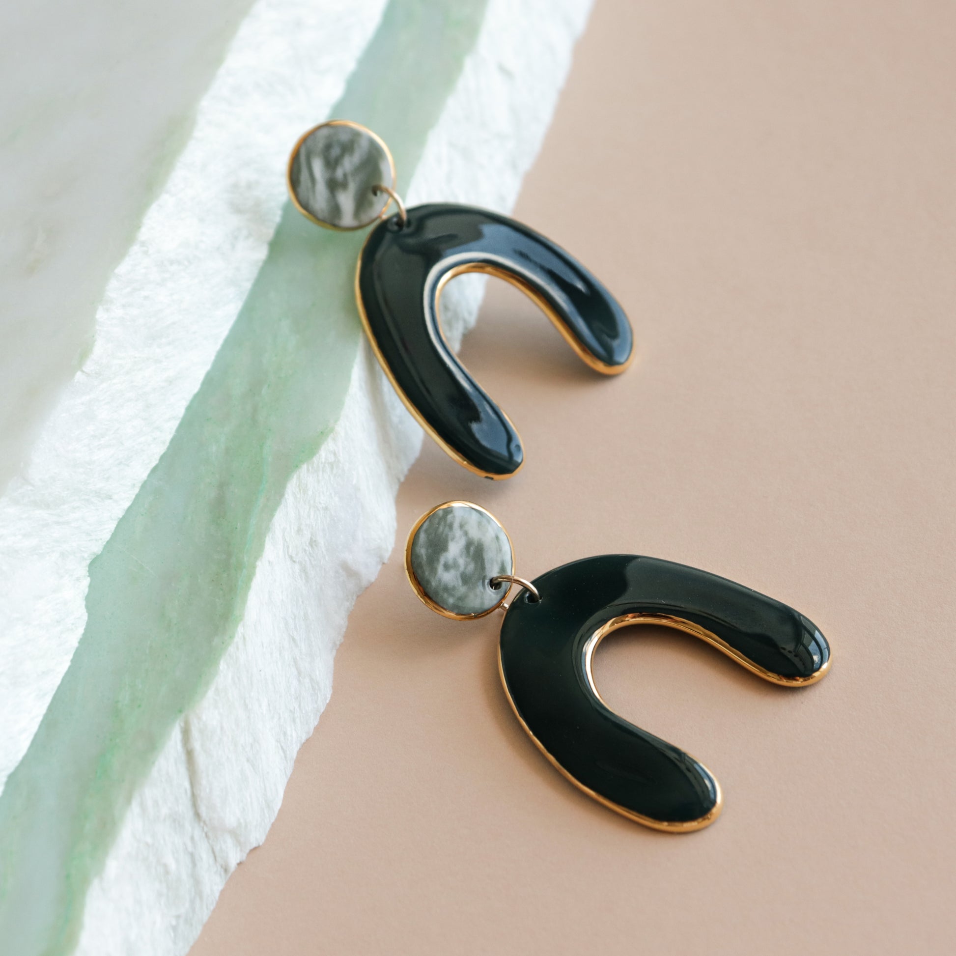 The Arquus Earrings by Rozenthal Jewelry are modern black and gold porcelain statement earrings with marbled circular studs, displayed on a textured white and beige surface.