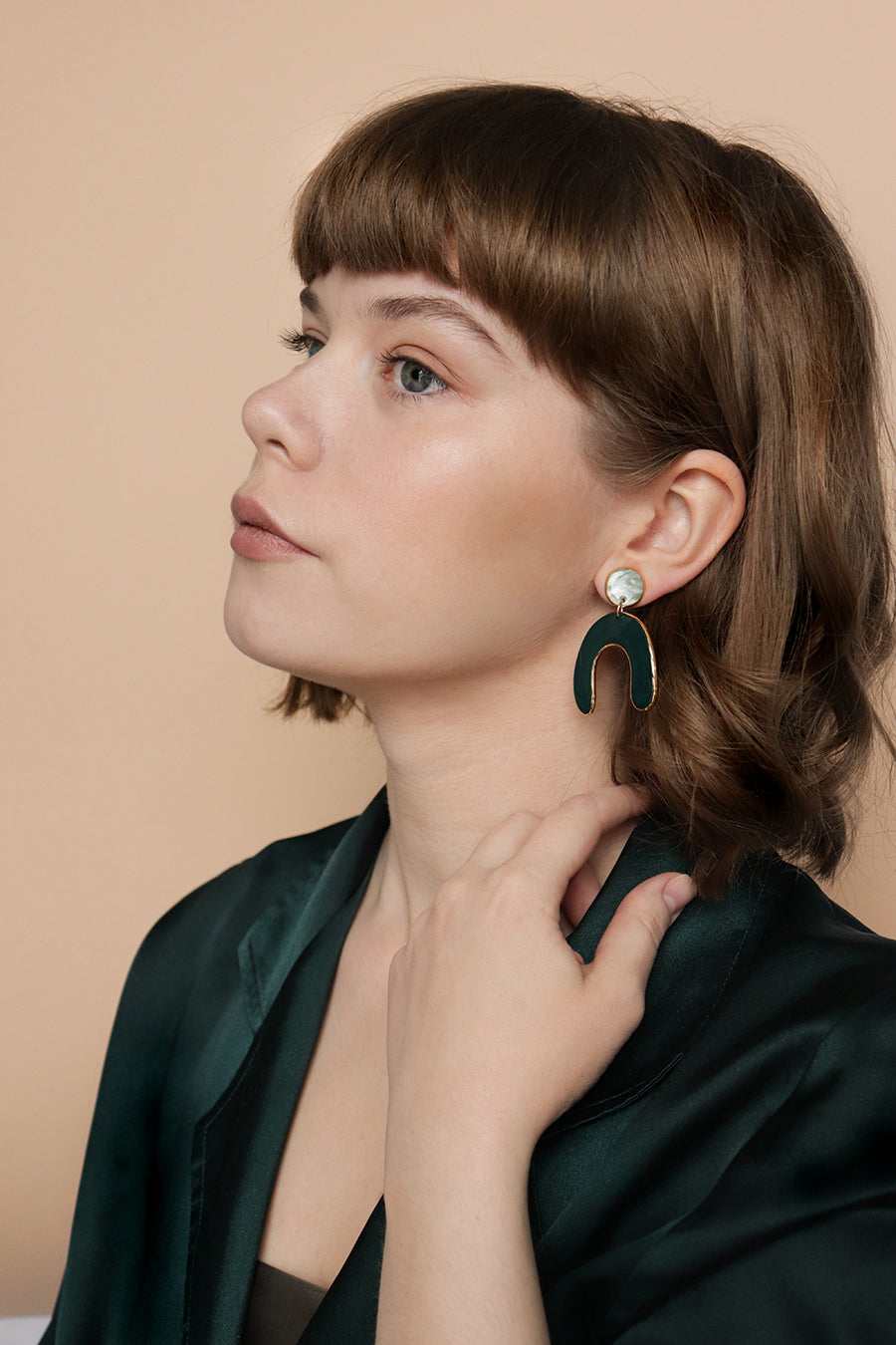 A woman with short brown hair and bangs wears a dark green satin top and the Arquus Earrings by Rozenthal Jewelry. She gazes upward with a neutral expression, touching her neck with one hand against a beige background.