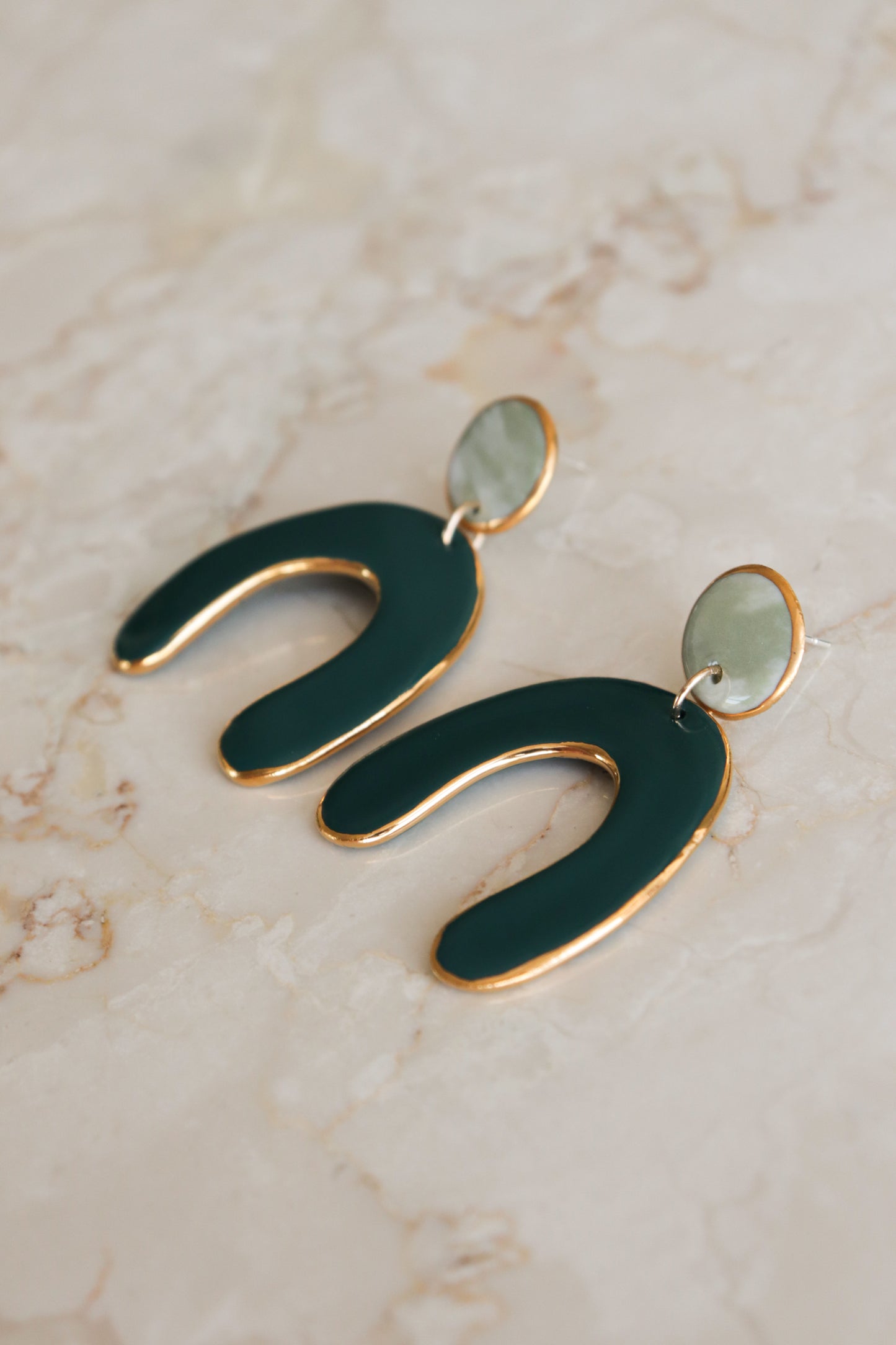 The Arquus Earrings by Rozenthal Jewelry feature modern green, arch-shaped porcelain pendants outlined in 24k gold, attached to small round light green studs, and are displayed on a light marble surface.